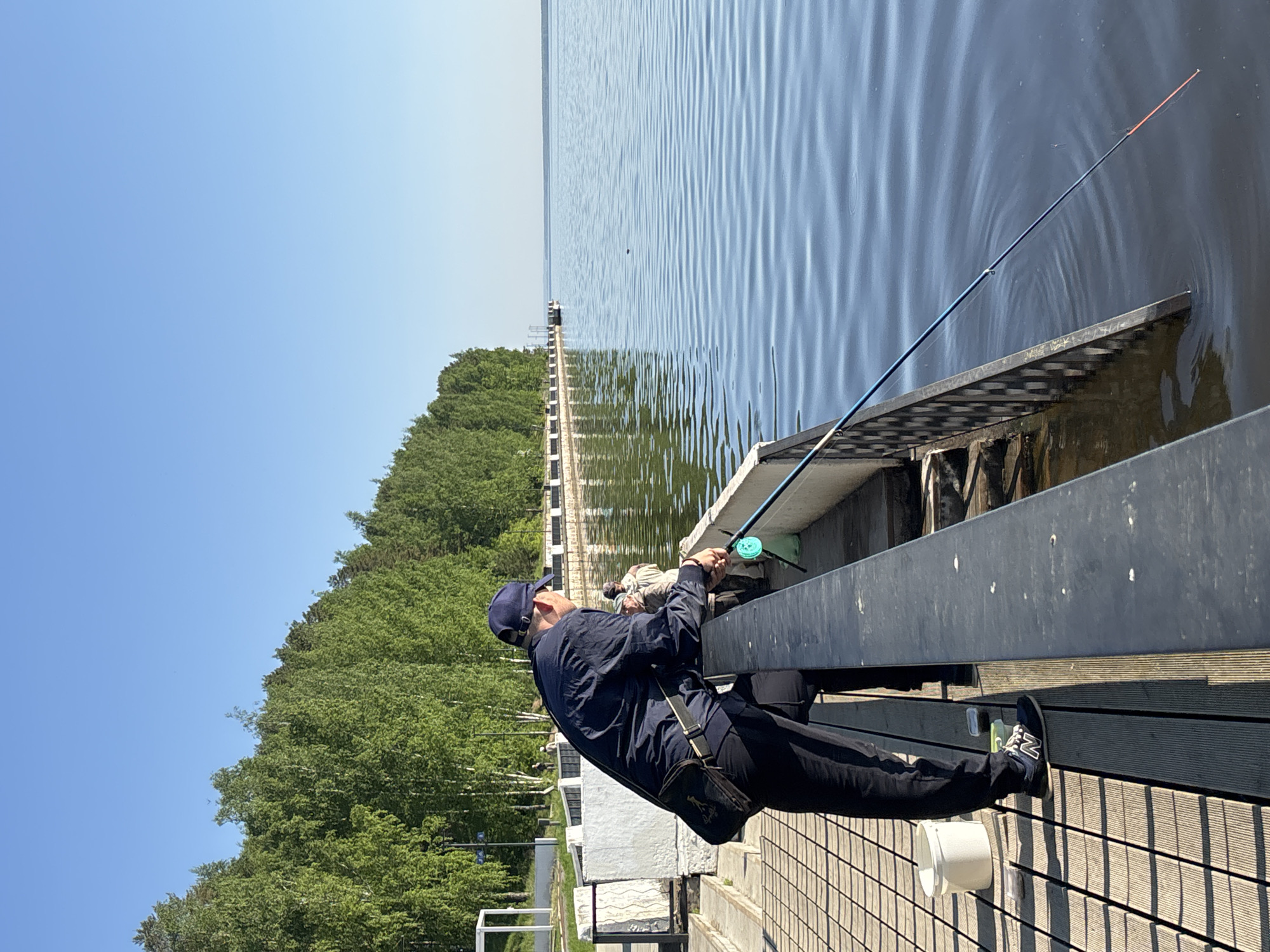 A man is fishing from a pier on the Kama River in the city of Chaikovsky, Perm Krai, Russia. He is wearing a dark blue jacket, dark pants, and a baseball cap. A fishing rod extends out over the water. A small white bucket is on the wooden pier next to his feet, and a dark bag is slung across his body. A long pier stretches out into the water, with trees along the shoreline to the left. The sky is clear and blue.