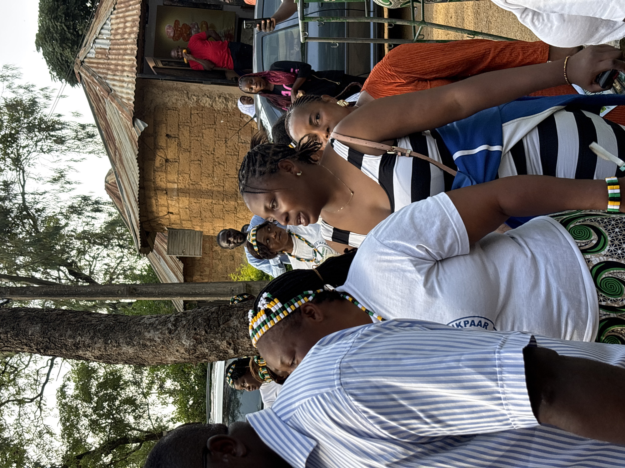In Jos, Nigeria, a group of people are gathered outdoors, seemingly at a cultural event or celebration. In the foreground, a man in a blue and white striped shirt stands to the left, partially obscuring a woman wearing a white t-shirt with a circular blue emblem and a black and white striped dress. She also wears a headband and necklace made of colorful beads. To her right, a woman in an orange long-sleeved shirt stands behind her. In the middle ground, several women with beaded headbands are visible, some in white t-shirts. Behind them, a man in a red shirt stands near a building with a corrugated metal roof, holding a piece of corn. The background includes trees and a car. The overall impression is one of a lively community gathering in a semi-urban or rural setting.