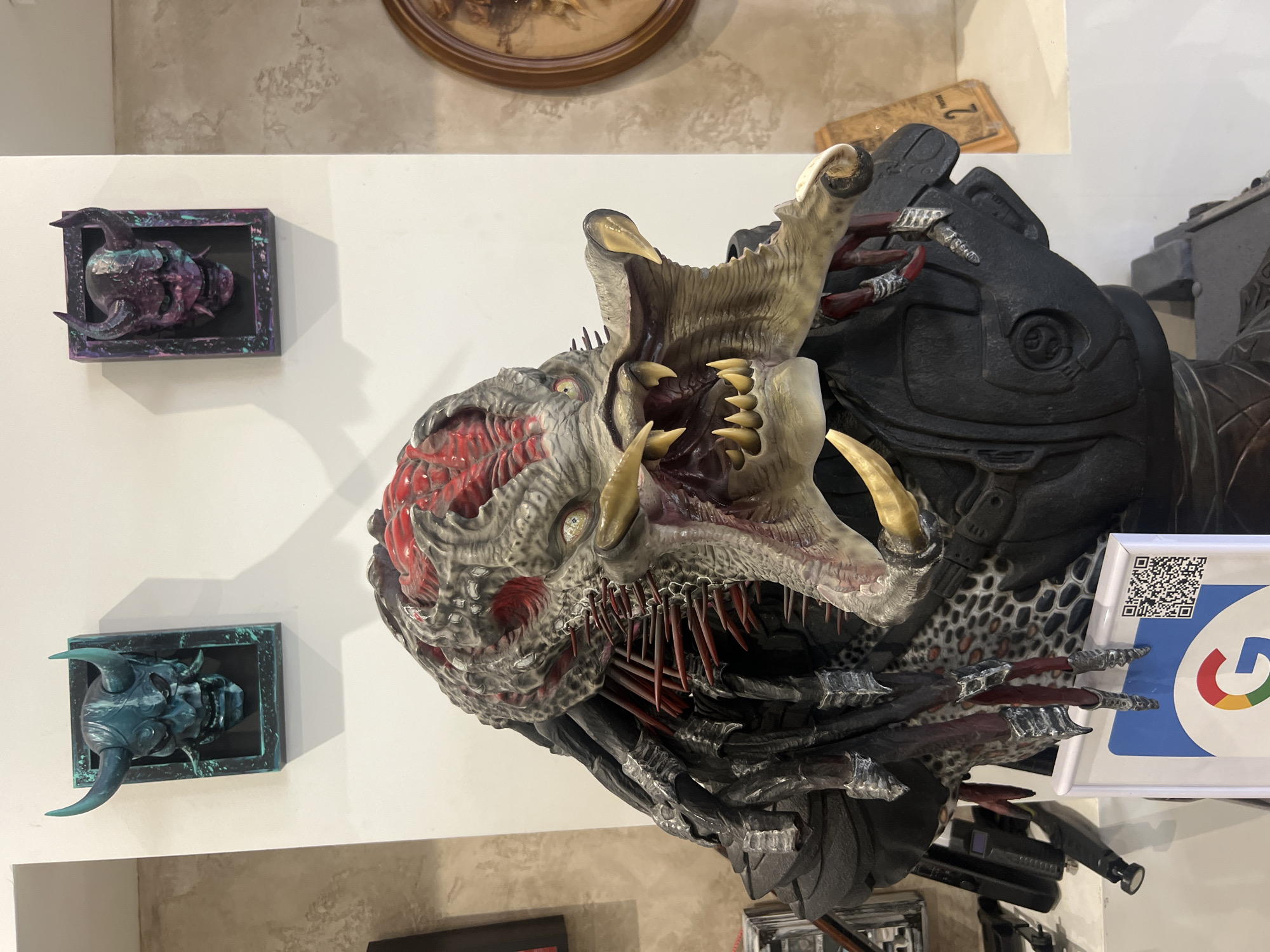 The image depicts a detailed, monstrous sculpture mounted on a wall, likely within an interior space. The sculpture features a creature with an open mouth, sharp teeth, and exposed, fleshy areas colored in red and gray. It is attached to a black, metallic structure resembling armor or biomechanical components. To the left of the sculpture are two smaller framed pieces, each containing a stylized head, one in shades of purple and the other in teal. A Google QR code sign is positioned near the sculpture. The scene suggests a display or exhibit, potentially within a gallery, museum, or themed entertainment location. The location is within the city of Sofia, Bulgaria.