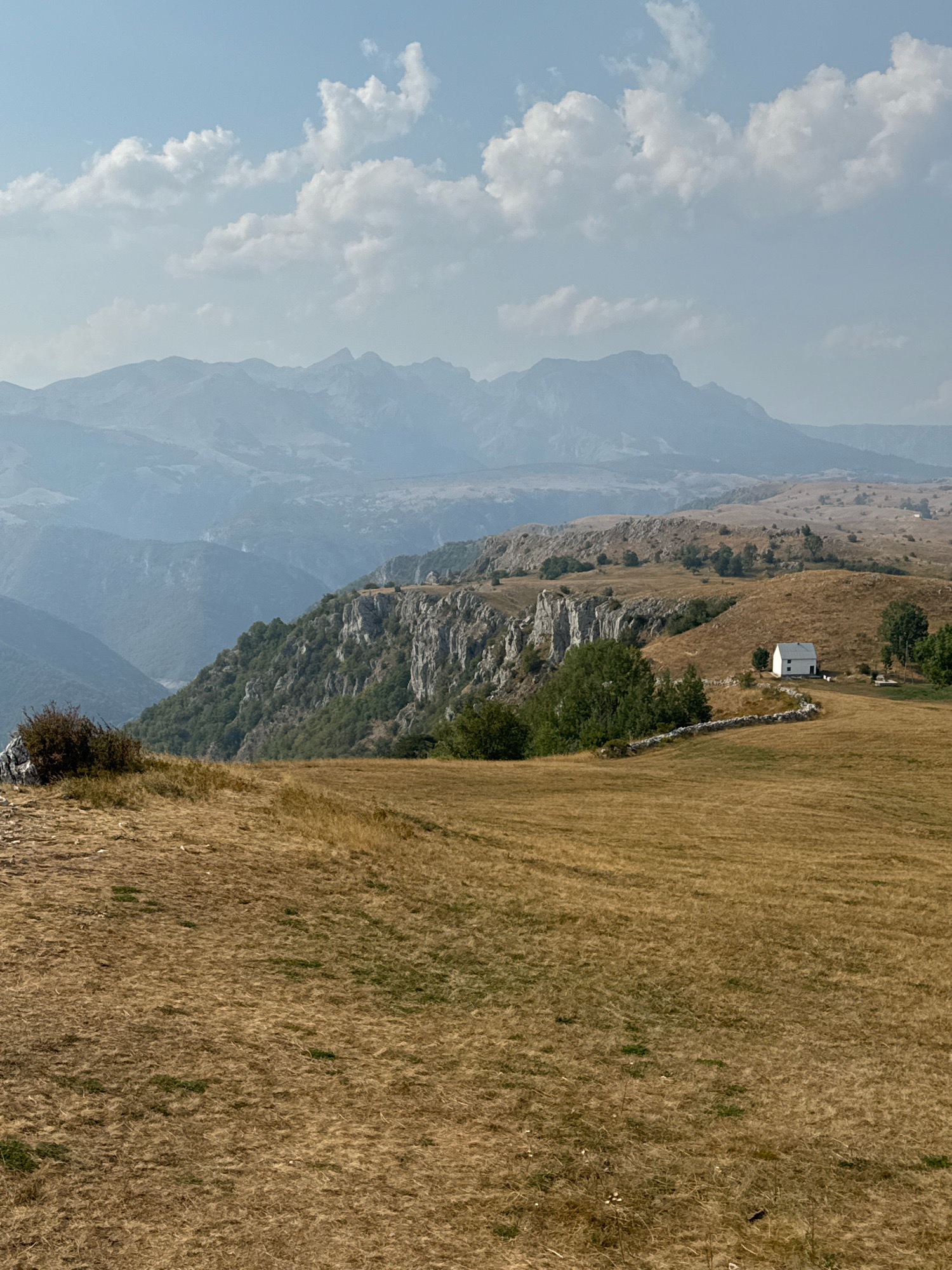The image depicts a mountainous landscape in the vicinity of Pišče, Montenegro. The foreground is dominated by dry, golden-brown grass covering rolling hills. In the midground, a steep, rocky cliff face covered in green vegetation descends into a valley. A small, white house with a dark roof is situated on a grassy knoll to the right of the cliff, bordered by a low, stone wall. Several trees, predominantly green, are scattered across the landscape. The background features layers of misty blue mountains stretching towards a hazy horizon, under a sky with scattered white clouds. The scene appears to be bathed in natural daylight, suggesting it was taken during the day. There are no people or animals visible in the image. The overall impression is one of rural tranquility and natural beauty.