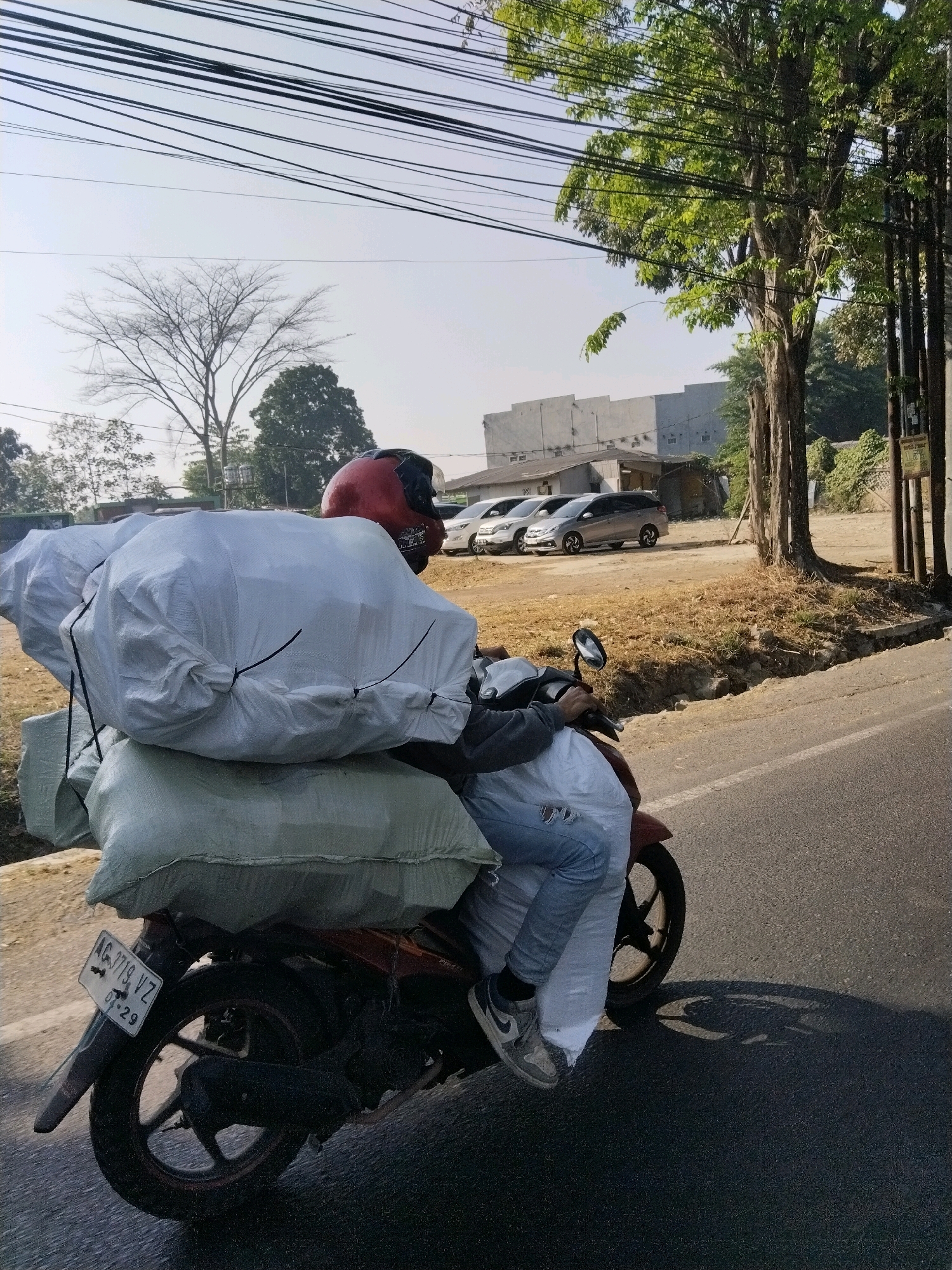 A person wearing a red helmet rides a red motorcycle loaded with large white and green bundles, down a paved road in Malang, Indonesia. The motorcycle's license plate reads "AG 1719 VZ 07-29". The person is wearing blue jeans with rips and gray sneakers. In the background, there are parked cars, buildings, trees, and overhead power lines. The scene is illuminated by sunlight, casting shadows on the road.