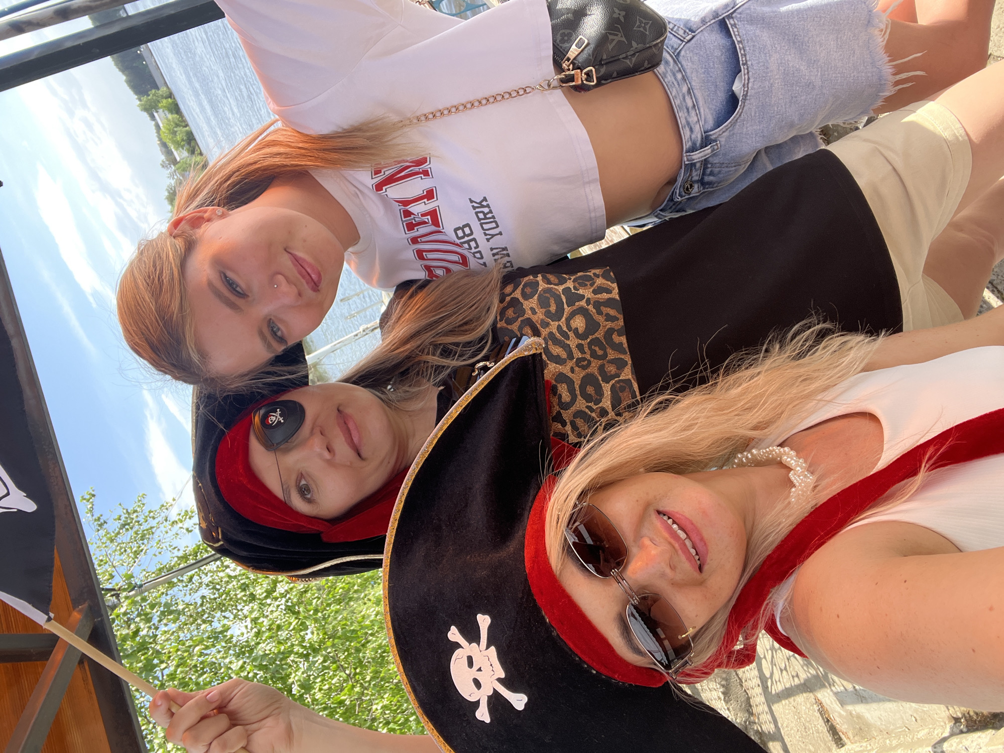 Three women pose for a selfie outdoors near the Kama River in Chaikovsky, Russia. One wears a white crop top with "Brooklyn New York" printed on it and light-wash denim shorts, with a small black purse on a gold chain. Two women wear pirate costumes with black hats and red accents; one has a skull and crossbones on the hat, sunglasses, and a pearl necklace. The other wears a patch over one eye and holds a small pirate flag. The background includes the Kama River, trees, and a partial view of the sky.