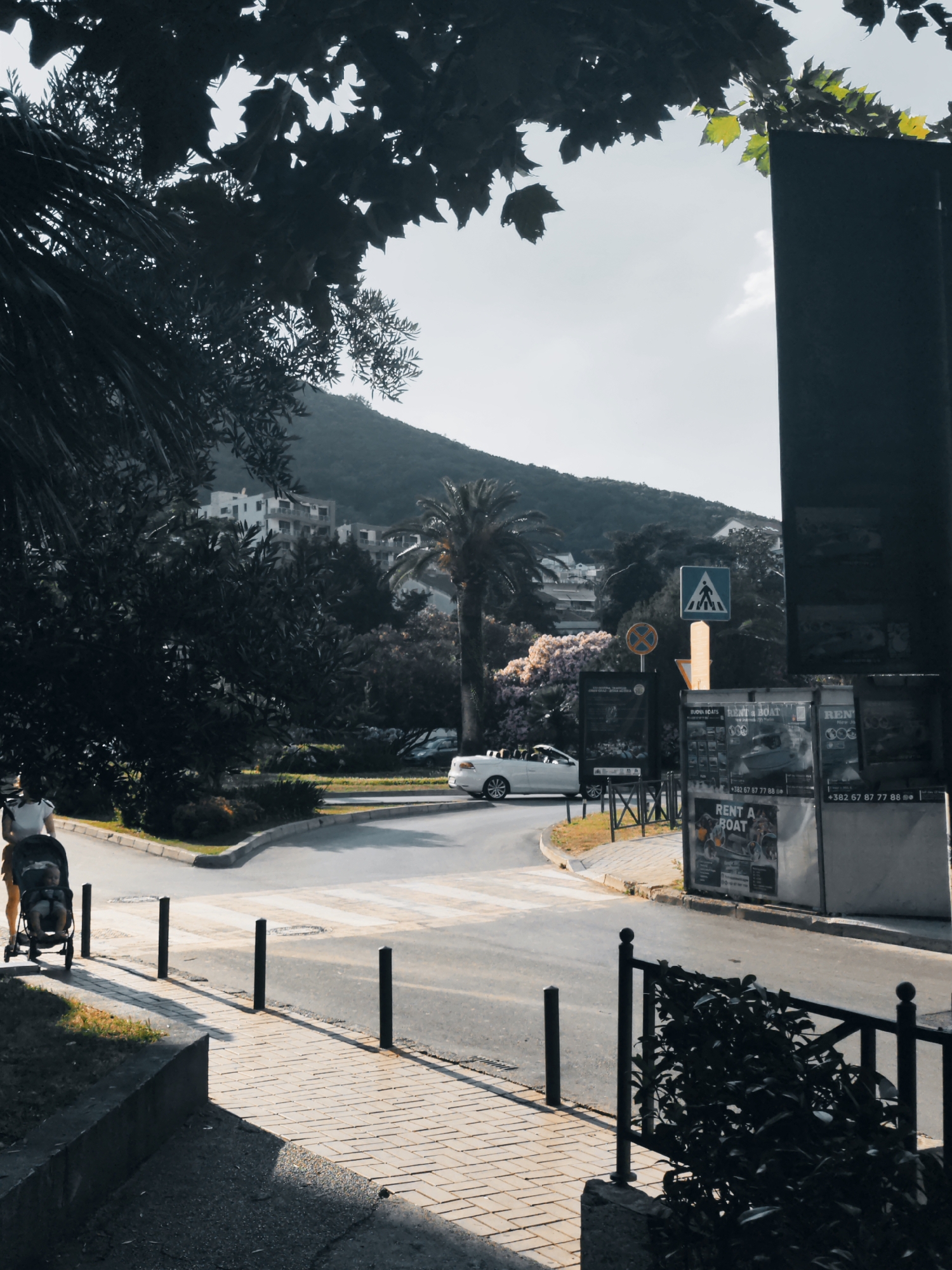 The image depicts a street scene in Budva, Montenegro. In the foreground, a person is pushing a stroller along a paved sidewalk. The stroller contains a baby. A pedestrian crossing is visible on the road ahead, with a white convertible car approaching it from the right. To the right of the road, there is a tall, dark structure displaying advertisements for "RENT A BOAT" services, with phone numbers clearly visible. A pedestrian crossing sign is mounted on a yellow pole. In the background, there are buildings and lush green trees, with a hazy mountain range in the far distance under a bright sky. The overall impression is of a sunny day in a coastal town.