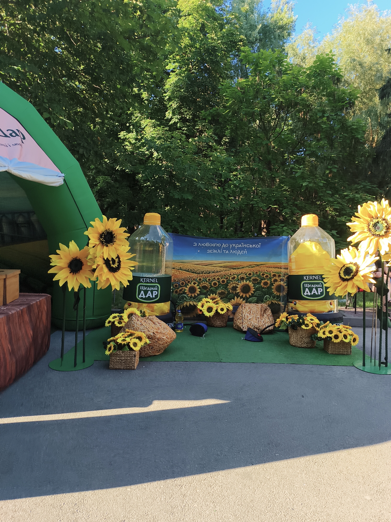 The image shows an outdoor event setup, likely a promotional booth, with a large inflatable green structure on the left, partially obscured by trees. The main display features two oversized bottles of what appears to be sunflower oil, each with the brand "KERNEL" and "Щедрий ДАР" visible. One bottle is a vibrant yellow, suggesting the oil's color, while the other is clear and filled with a yellow substance. In front of the bottles are various decorative elements, including artificial sunflowers arranged in baskets and on stands, as well as woven decorative items. A banner in the background depicts a field of sunflowers with Ukrainian text that translates to "With love to Ukrainian land and people." The ground is paved, with a green artificial turf area for the display. The overall setting is outdoors, with lush green trees and a clear blue sky visible in the background, suggesting a sunny day. The event appears to be in Ternopil, Ukraine, given the presence of the brand and Ukrainian text. There are no people clearly visible in the image.