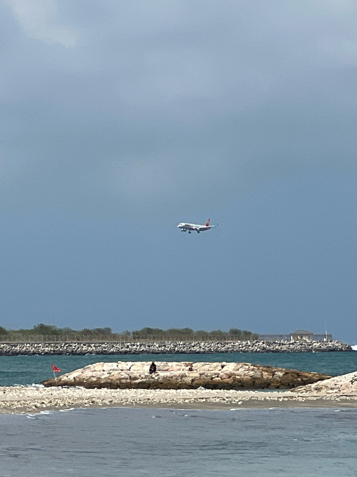 An airplane is shown in flight against a blue, overcast sky. The airplane is white with red accents and appears to be descending, possibly for landing. Below the sky, a body of water is visible, with waves lapping at a rocky shore. A small, rocky outcrop extends into the water from the left side of the frame, and a person is seated on this outcrop. Further in the background, a long, stone breakwater stretches across the horizon, with trees and structures visible behind it. The scene is calm and serene, with the airplane as the most dynamic element. This scene is not located in Kuta, Indonesia.