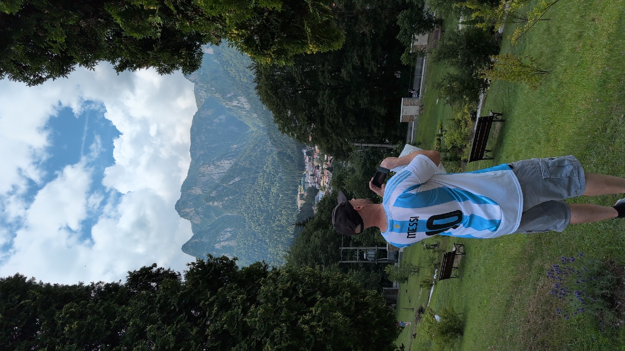 A man wearing a white and light blue striped jersey with "MESSI 10" on the back stands in a grassy area, holding a phone in his hands as if taking a picture. He is facing away from the camera and towards a mountainous landscape. In the foreground, there is a small lavender plant. In the background, there are trees, benches, and a town nestled at the base of the mountains. The sky is cloudy and blue. The overall scene suggests a tourist enjoying the scenery in Bușteni, Romania.