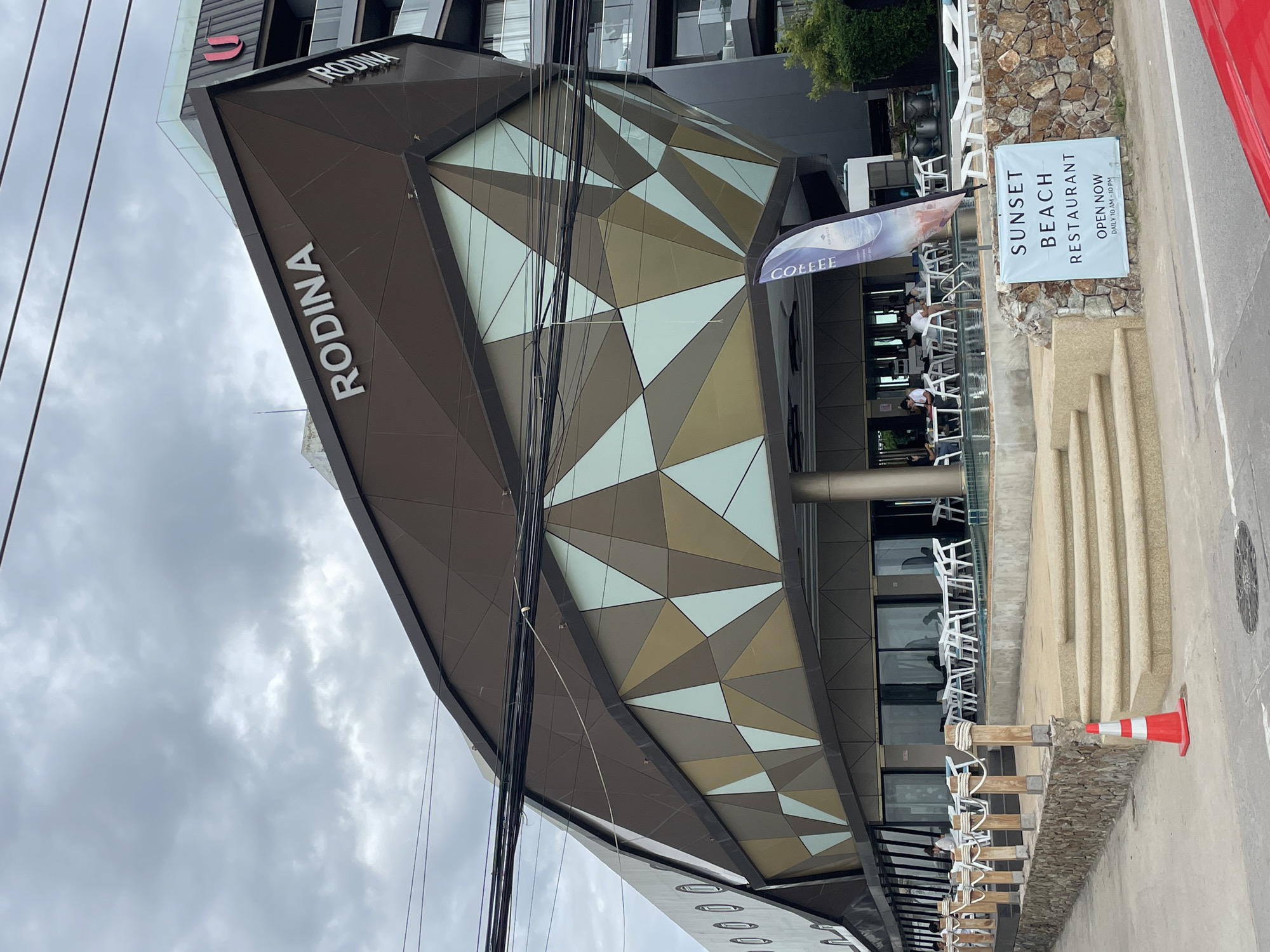 This is a medium-angle shot of the Rodina building in Pattaya City, Thailand, on an overcast day. The building has a modern, angular design with a facade featuring geometric patterns in shades of brown, gold, and light blue. The word "RODINA" is prominently displayed in white lettering on the upper part of the building. In front of the building, there is an outdoor seating area with white tables and chairs, some set up around a pool. A blue and white banner with the word "COFFEE" is visible near the entrance. A sign on a stone wall reads "SUNSET BEACH RESTAURANT OPEN NOW DAILY 10 AM - 10 PM". Several people are seated at the tables, some appearing to be dining or socializing. A red traffic cone is on the pavement in the foreground, and power lines are visible in the sky above the building. The overall impression is of a commercial establishment, possibly a hotel or resort with a restaurant.
