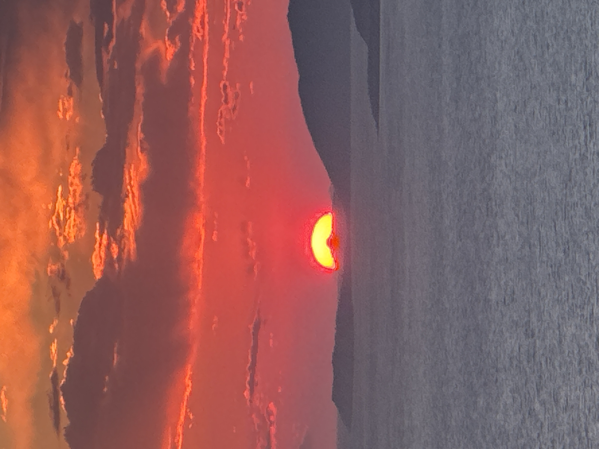 This image captures a vibrant sunset over the sea, with the sun partially submerged behind distant, dark hills. The sky is ablaze with intense shades of red and orange, illuminated by the setting sun. Wispy clouds are scattered across the sky, catching the fiery hues of the sunset. The sea below is a muted grey, reflecting the dramatic colors of the sky in a less intense manner. The scene appears to be a coastal landscape, possibly the coast of Zagora, Montenegro, given the geographical context provided. There are no people or distinct objects visible in the image, focusing purely on the natural spectacle of the sunset and its effect on the environment. The lighting is dramatic and diffused, characteristic of twilight.