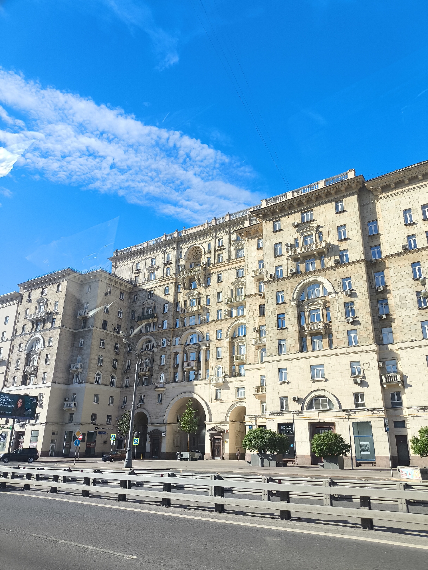 The image captures a grand, pale-yellow apartment building with arched entrances and ornate balconies, indicative of a Stalinist Empire style architecture. The building is bathed in sunlight, suggesting it is daytime. A bright blue sky with scattered white clouds forms the backdrop.
In the foreground, a multi-lane road runs parallel to the building, separated by a metallic guardrail. The road appears to be an urban street or highway. A few cars are visible on the road and a pedestrian walkway in front of the building. There are also trees and streetlights along the walkway.
On the left side of the building, a large billboard displays an advertisement with text in Cyrillic, featuring a woman's face and the words "Стать учителем нового поколения" (Become a teacher of the new generation). Other signs with Cyrillic text are also visible on the building's ground floor, indicating commercial establishments.
The overall scene suggests a sunny day in a city, likely Eastern Europe given the Cyrillic text and architectural style. The building's imposing facade and the sunny weather create a sense of permanence and urban vibrancy. No specific activity or event is clearly depicted, but the presence of cars and pedestrian areas implies everyday urban life.