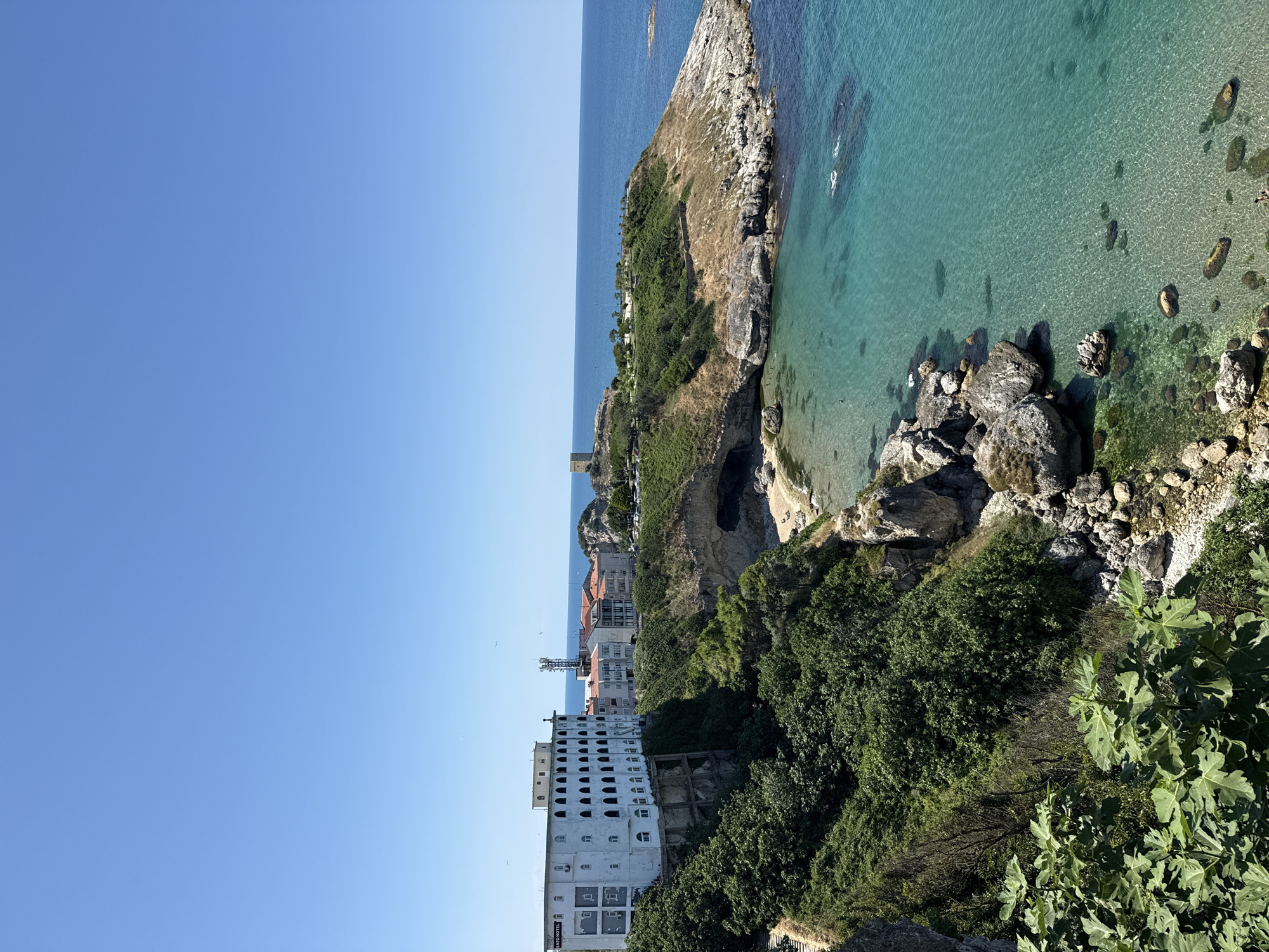 This is a medium-shot, high-angle, vertical photograph taken on a clear, sunny day, overlooking the coastal city of Şile, Turkey. The foreground features lush green foliage, likely fig leaves, partially obscuring a rocky incline leading down to the sea. The middle ground showcases a dramatic coastline with rocky cliffs, a small sandy cove accessible via a sea cave, and the turquoise waters of the Black Sea.
Dominating the left side of the middle ground is a large, white, multi-story hotel with numerous arched windows and balconies, identified by a sign as "SATO HOTEL". Behind the hotel, several other buildings with red-tiled roofs are visible, suggesting a developed urban area. Further along the coast, on a hilltop to the right, a medieval stone tower stands prominently, characteristic of historical fortifications. The sea stretches to the horizon, meeting a clear blue sky. A few birds are visible in the sky, and a handful of people can be seen swimming or wading in the shallow water near the shore. The overall scene depicts a picturesque coastal landscape combining natural beauty with architectural elements.
