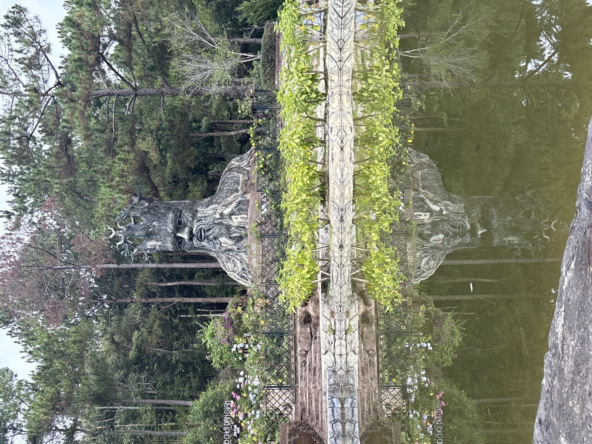 The image displays a serene outdoor scene, appearing to be a landscaped garden or park, rotated 90 degrees clockwise from its natural orientation. When viewed correctly, a large, weathered stone or concrete sculpture of a human-like face, possibly representing a "Green Man" or nature spirit, dominates the mid-ground on the left. The sculpture features foliage-like elements and a stern, ancient expression.
In front of the sculpture and extending across the lower half of the frame is a calm body of water, likely a pond or lake, offering clear reflections of the surrounding elements. The water's surface is still, mirroring the dense pine forest that forms the backdrop, as well as parts of the sculpture and adjacent vegetation.
Along the water's edge, a patterned stone pathway or low wall, possibly part of a decorative railing system, stretches horizontally. Small green plants grow on and around this structure. Beyond this, a variety of colorful flowering plants in shades of purple, pink, white, and yellow are visible, indicating cultivated garden beds. Intricate black metal fencing or railings are also present, separating different sections of the garden.
The setting appears peaceful and natural, with a rich green palette from the numerous trees and plants. The lighting is soft and diffused, suggesting an overcast day or a time of day with indirect sunlight, contributing to the tranquil atmosphere. There are no people visible, and no specific activity is taking place other than the static display of the garden and sculpture.
Visible text on a small white sign or banner in the lower right (when correctly oriented) reads "ĐỒ CHƠI DÂN GIAN", which translates to "FOLK TOYS". Another partial word, possibly "với", is faintly discernible above it.