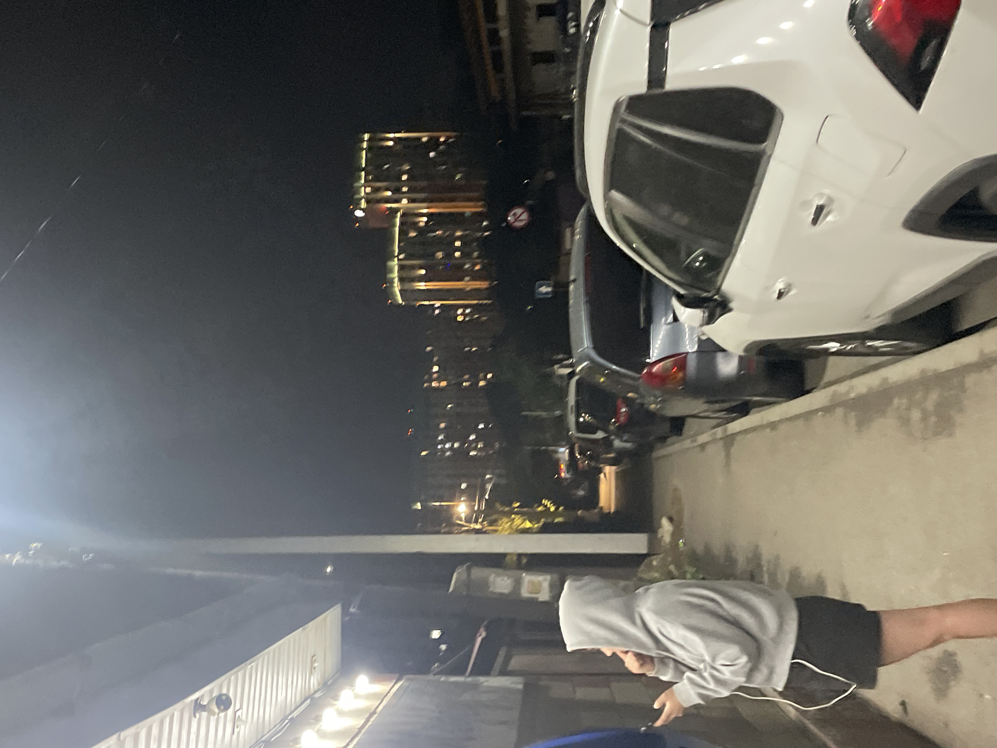 A young person in a grey hooded sweatshirt and black shorts stands on a sidewalk at night, looking at a phone. They are wearing earbuds. The sidewalk is lined with parked cars, including a white SUV in the foreground. In the background, apartment buildings with illuminated windows are visible. The scene appears to be in Almaty, Kazakhstan.
