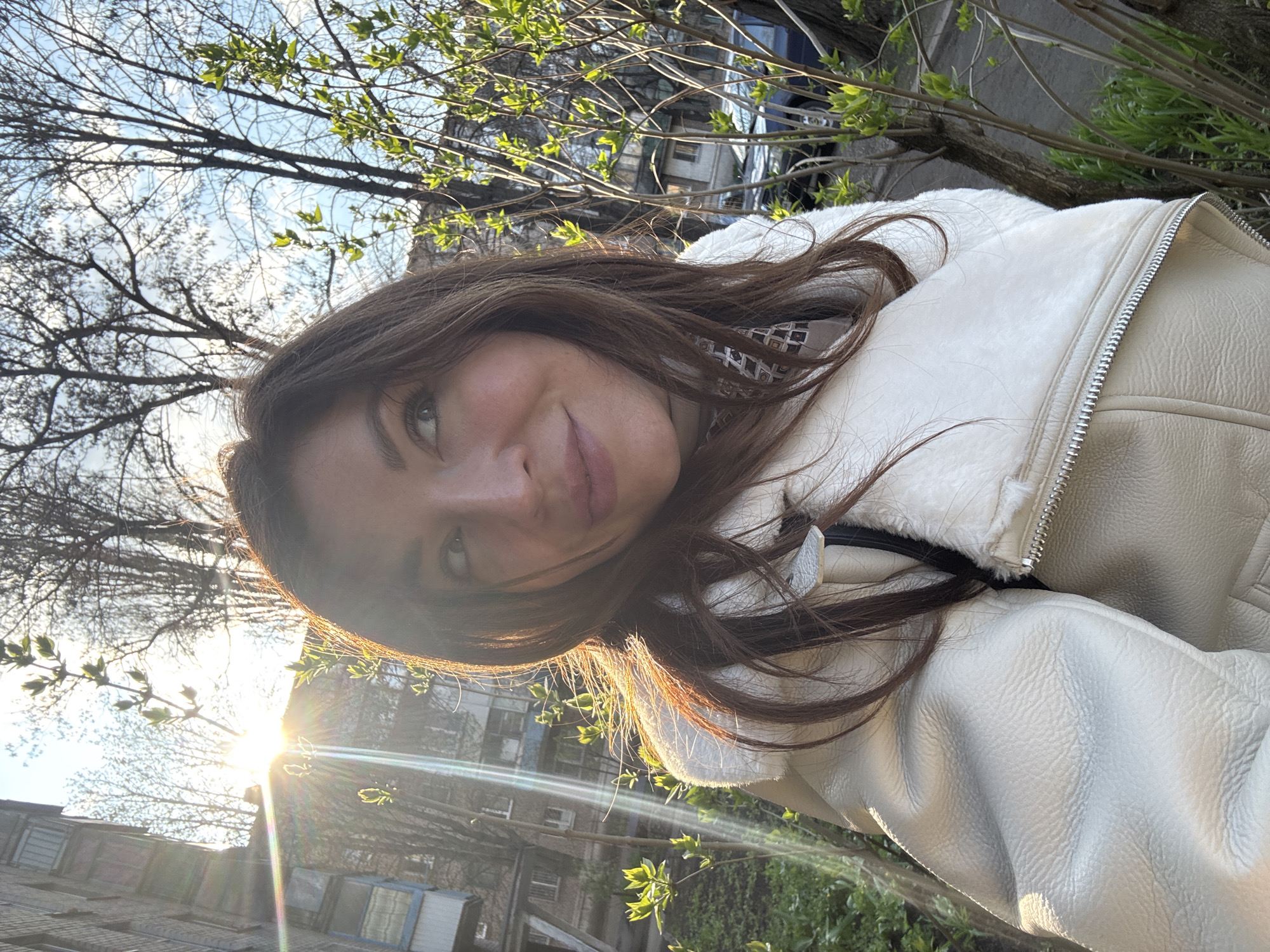 A medium shot captures a young woman outdoors in an urban setting. She is turned slightly to her right, facing the camera with a soft smile. She wears a light-colored jacket with a fluffy collar. The background features trees with new spring foliage, and a city building is faintly visible with a sunburst. The lighting appears natural, possibly late afternoon or early morning, casting a warm glow on her face and hair.