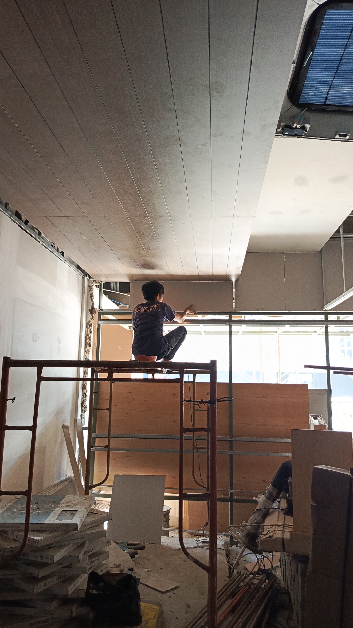 A person is working on installing a ceiling indoors. The person is wearing a dark blue t-shirt and dark pants, and is crouched on scaffolding. They appear to be attaching a ceiling panel. The ceiling is composed of light brown wood panels. The area appears to be under construction, with exposed framing and scaffolding. There are stacks of building materials on the floor. The lighting suggests daytime, with natural light coming from windows. This scene is located in Tangerang, Indonesia.