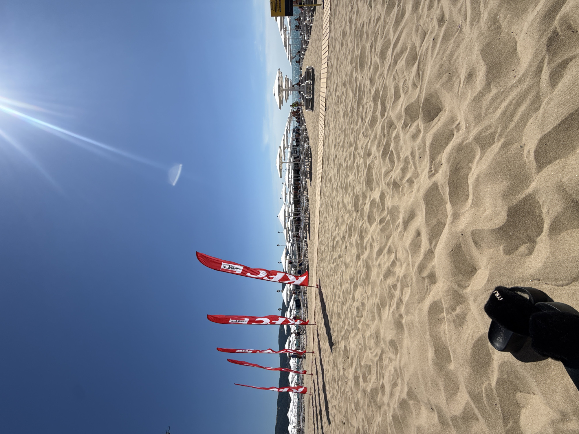 The image depicts a sunny day at a sandy beach in Nesebar, Bulgaria. The foreground features ripples of sand, with a pair of black furry slides placed on the sand. In the midground, a row of red flags with white logos, appearing to be KFC branding, stands vertically. Beyond the flags, rows of white beach umbrellas and lounge chairs are set up on the sand. The background shows the blue sea under a clear, bright blue sky with the sun emitting rays. A few people are visible in the distance, seated or walking among the beach amenities. A wooden walkway runs horizontally across the sandy area, leading towards the umbrellas.