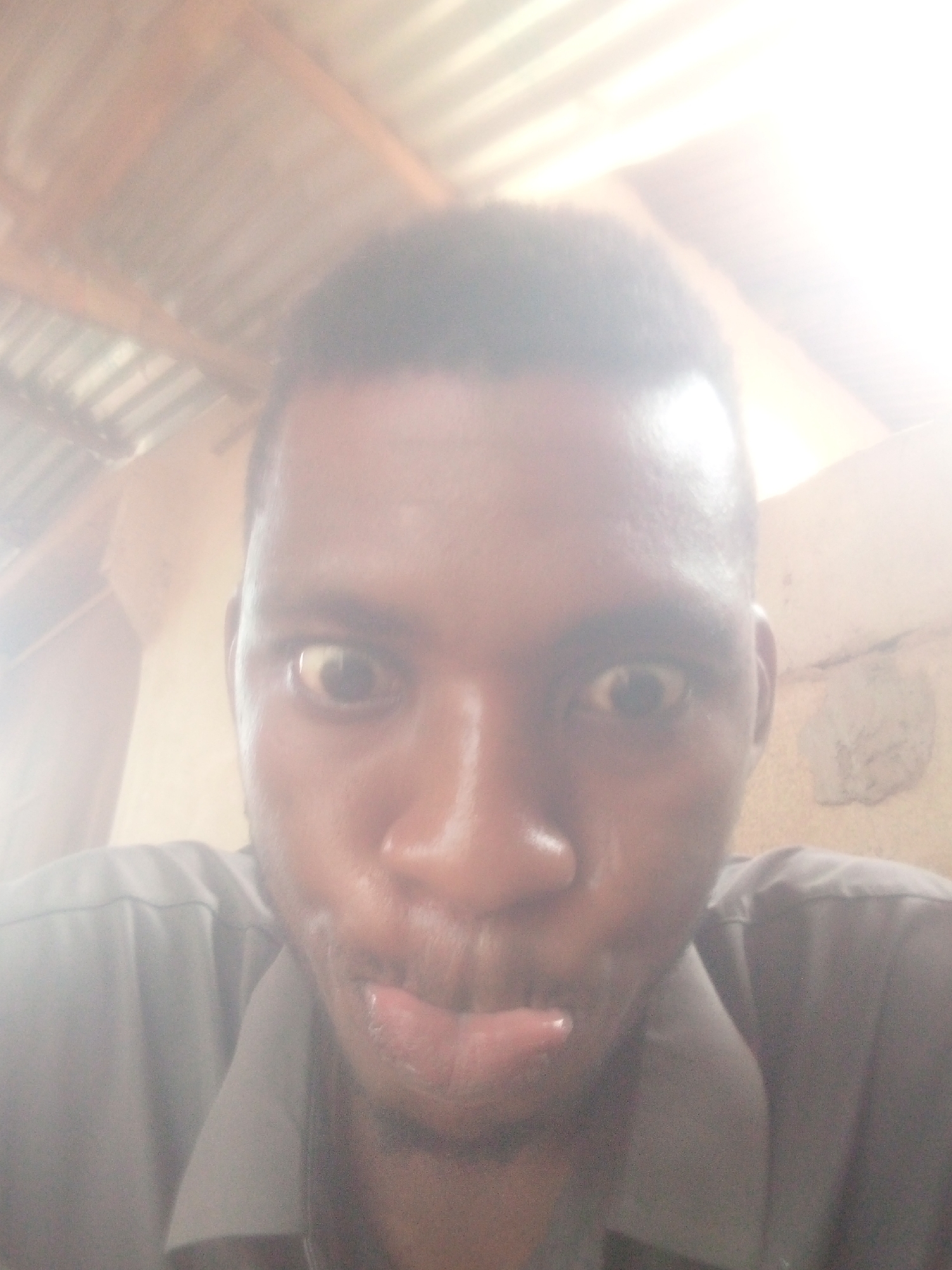 A close-up, brightly lit selfie captures a person, likely male, with a playful expression. The individual has dark skin and short, dark hair, and is looking directly at the camera with wide, slightly surprised eyes. Their tongue is subtly sticking out, suggesting a humorous or mischievous mood. The person is wearing a dark-colored, collared shirt.
The background is significantly overexposed and blurred, particularly towards the upper right, indicating a strong light source, possibly direct sunlight or a bright window. Despite the overexposure, hints of the setting are discernible: the upper left portion of the image reveals what appears to be a corrugated metal roof or ceiling structure with visible wooden support beams. The right side shows an indistinct wall with some texture, potentially concrete or mud brick. The overall image has a washed-out, high-key aesthetic due to the intense lighting. No visible text is present.