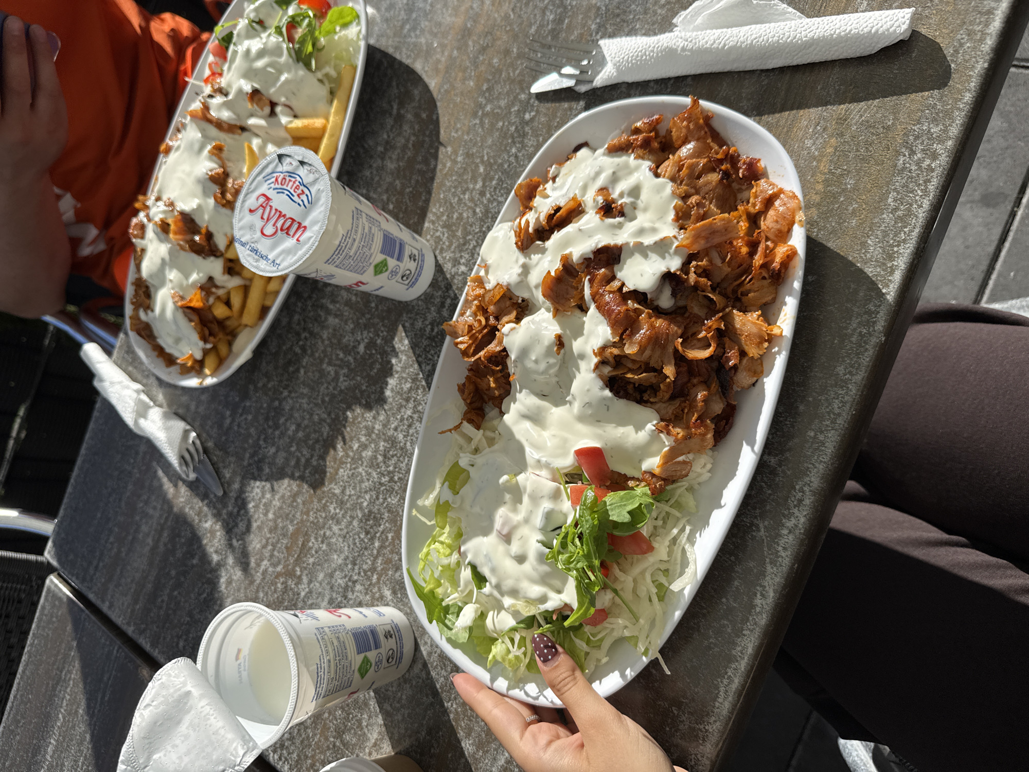 The image shows two plates of Döner meat, topped with a white sauce, served on a bed of shredded lettuce and tomatoes, along with a side of french fries on one of the plates. Two containers of Ayran are present. One plate is being held by a hand with polished nails. The setting appears to be an outdoor table. Given the presence of Ayran and Döner, the location could be a restaurant in Bruchköbel, Germany.