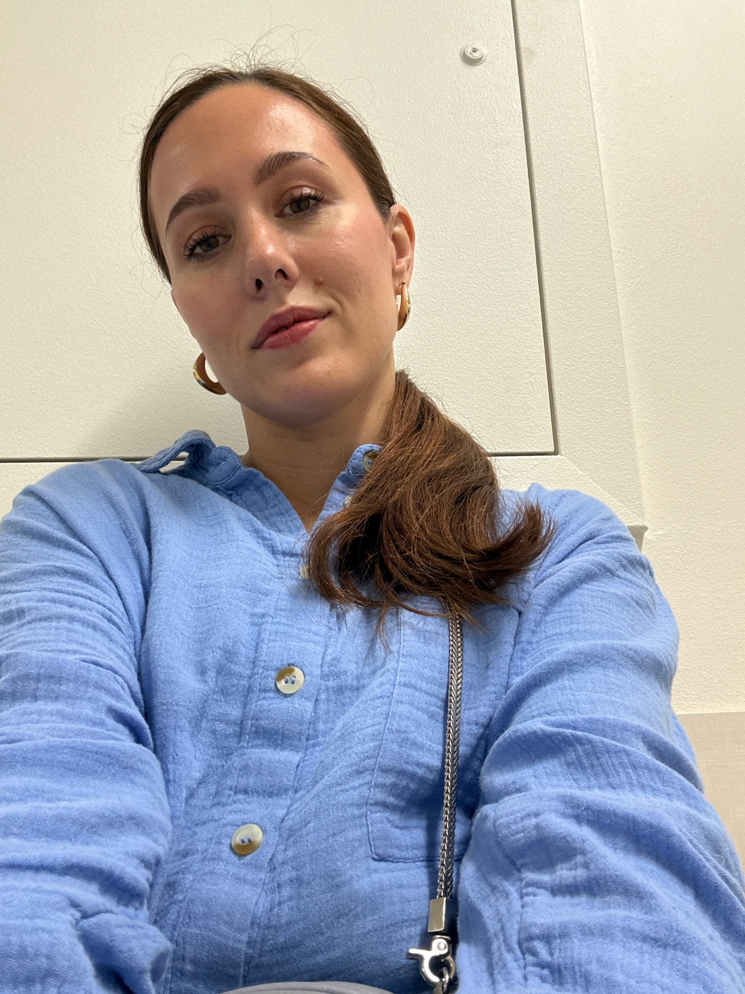 A close-up, overhead selfie features a woman with medium-toned skin and brown hair pulled back into a ponytail. She is wearing a light blue, long-sleeved, collared shirt with buttons. She has on large gold hoop earrings. Her makeup includes eyeshadow, eyeliner, and lip gloss. The background appears to be an indoor wall, possibly in a building in Riga, Latvia, with a visible ceiling fixture and a seam in the wall. She is wearing a silver chain strap from a purse.