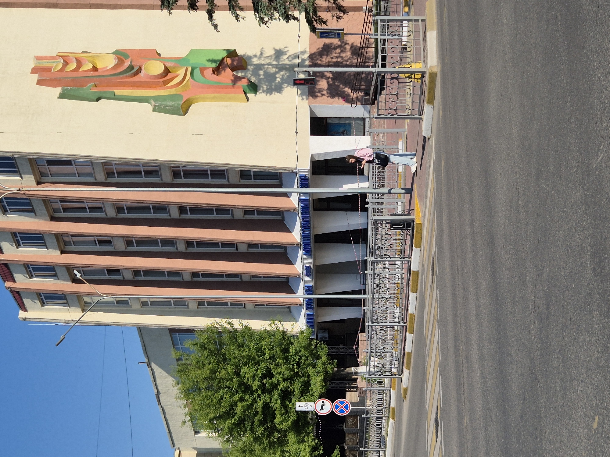 A tall building stands next to a road on a clear, sunny day in Kokshetau, Kazakhstan. The building has multiple stories with rows of windows and a large, colorful abstract artwork on its upper section. A woman wearing a pink shirt and jeans leans against a gray metal railing along the building's edge. The road, made of asphalt, shows zebra stripes of white paint in the foreground and extends into the distance. In the lower left corner, lush green trees surround two adjacent road signs.