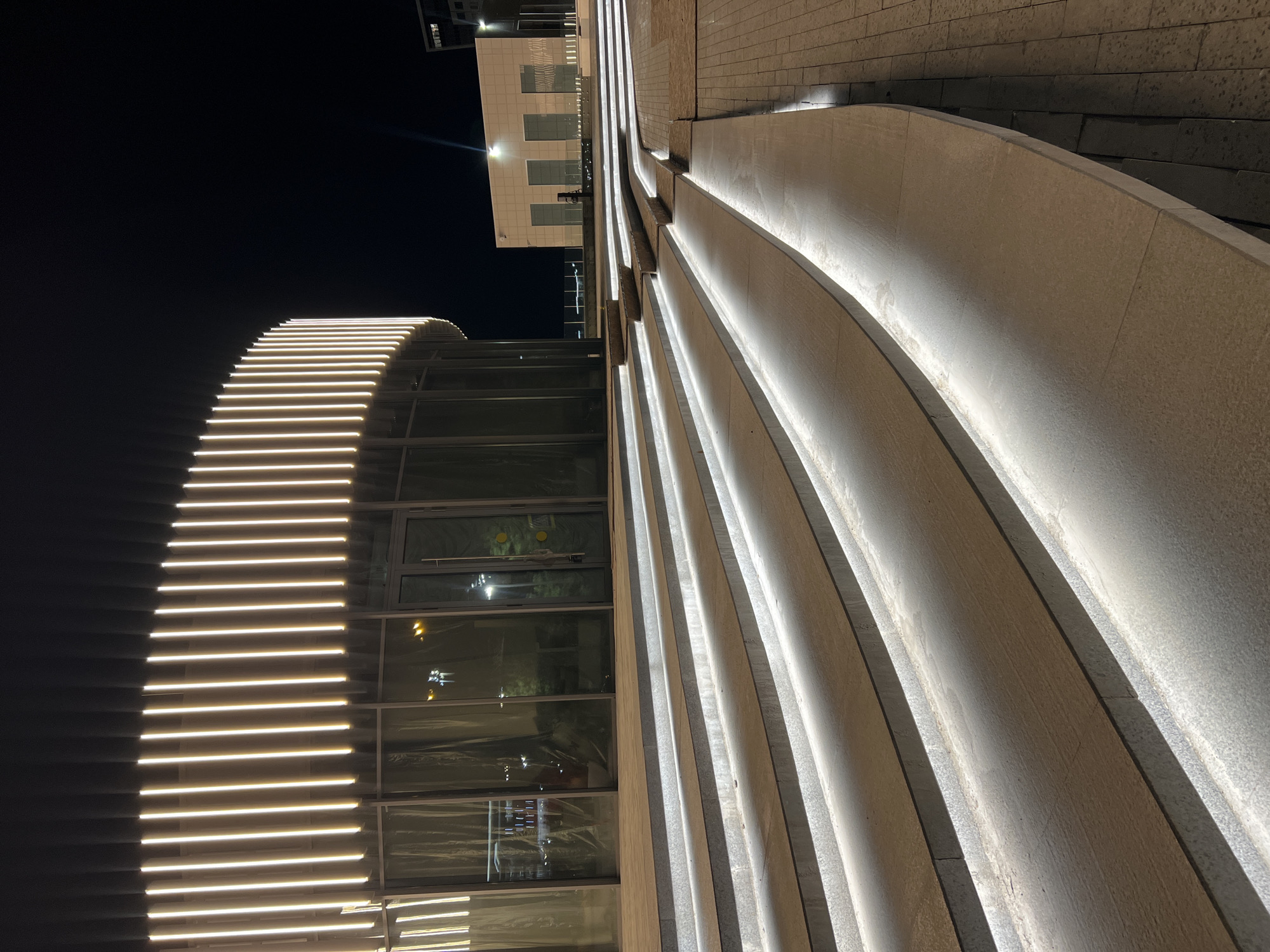 The image shows an exterior view of a building at night in Kazan, Russia. The foreground features a series of curved, lit architectural elements, possibly walls or barriers, with embedded light strips creating a linear illumination effect. The adjacent building section is modern, with large glass windows and corresponding light strips on the exterior. A portion of another building is visible in the upper background, also illuminated. The environment appears to be a well-lit, urban space with paved surfaces. There are no people visible in the image.