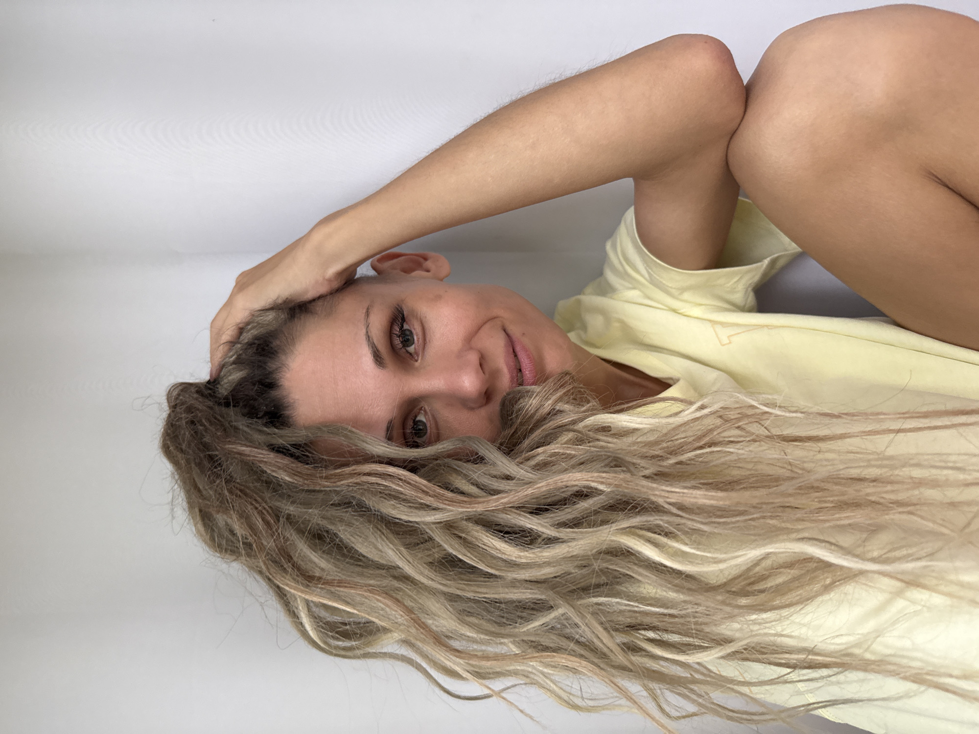 A medium shot captures a woman with long, wavy, blonde hair reaching over her left shoulder, resting her right hand on the top of her head. She is wearing a light yellow t-shirt and is positioned in the bottom right corner of the frame, facing left with her face slightly turned towards the camera. Her eyes are open, and she has a subtle smile. The background is a plain white wall, creating a minimalist setting. No specific location in Zhukovo, Russia is discernible from the image.