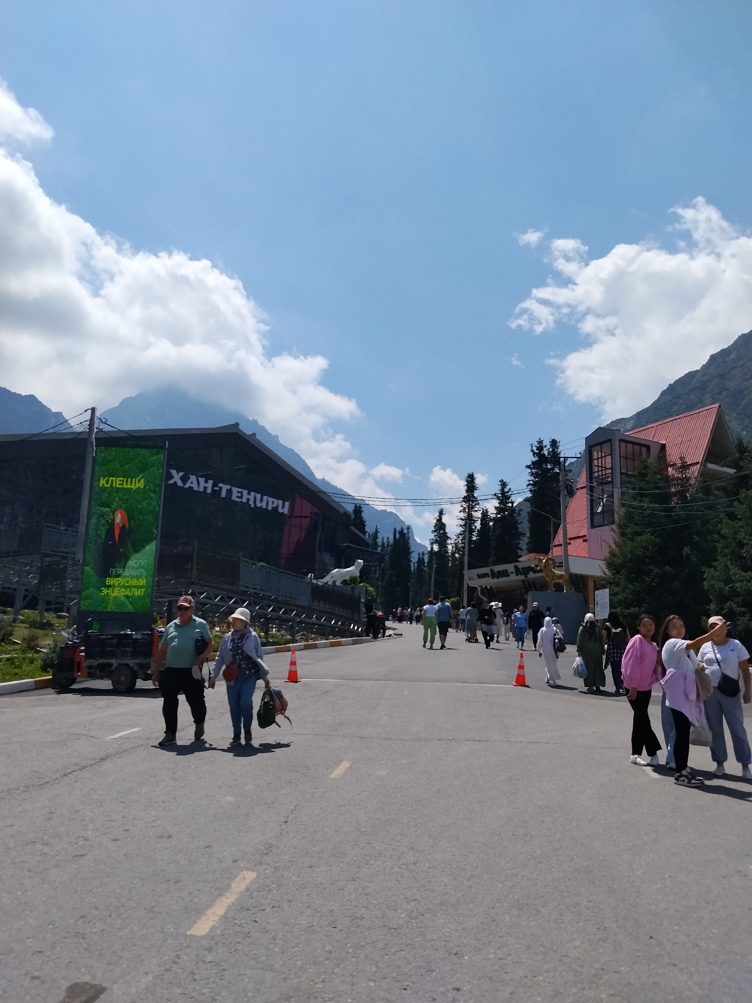 The image depicts a scenic outdoor scene in an mountainous area with a clear blue sky and scattered white clouds. The foreground features a paved road with yellow lane markings, where several people are walking. A couple, a man and a woman, are walking hand-in-hand in the lower left of the frame, while other individuals and groups are visible further down the road.
On the left side of the road, there is a large building with a prominent green banner displaying text and an image of a toucan. The banner appears to be an advertisement or informational display. To the right of this building, another building with red roofing and glass structures is visible, surrounded by numerous evergreen trees. A golden lion statue is perched on the roof of this building.
In the background, majestic mountains rise up, partially obscured by clouds. The overall setting suggests a tourist or recreational area, possibly a resort or a park, in a mountainous region. The presence of the buildings, signage, statues, and the flow of people indicates a developed area within a natural landscape. The image seems to be taken from the perspective of someone walking towards the buildings.