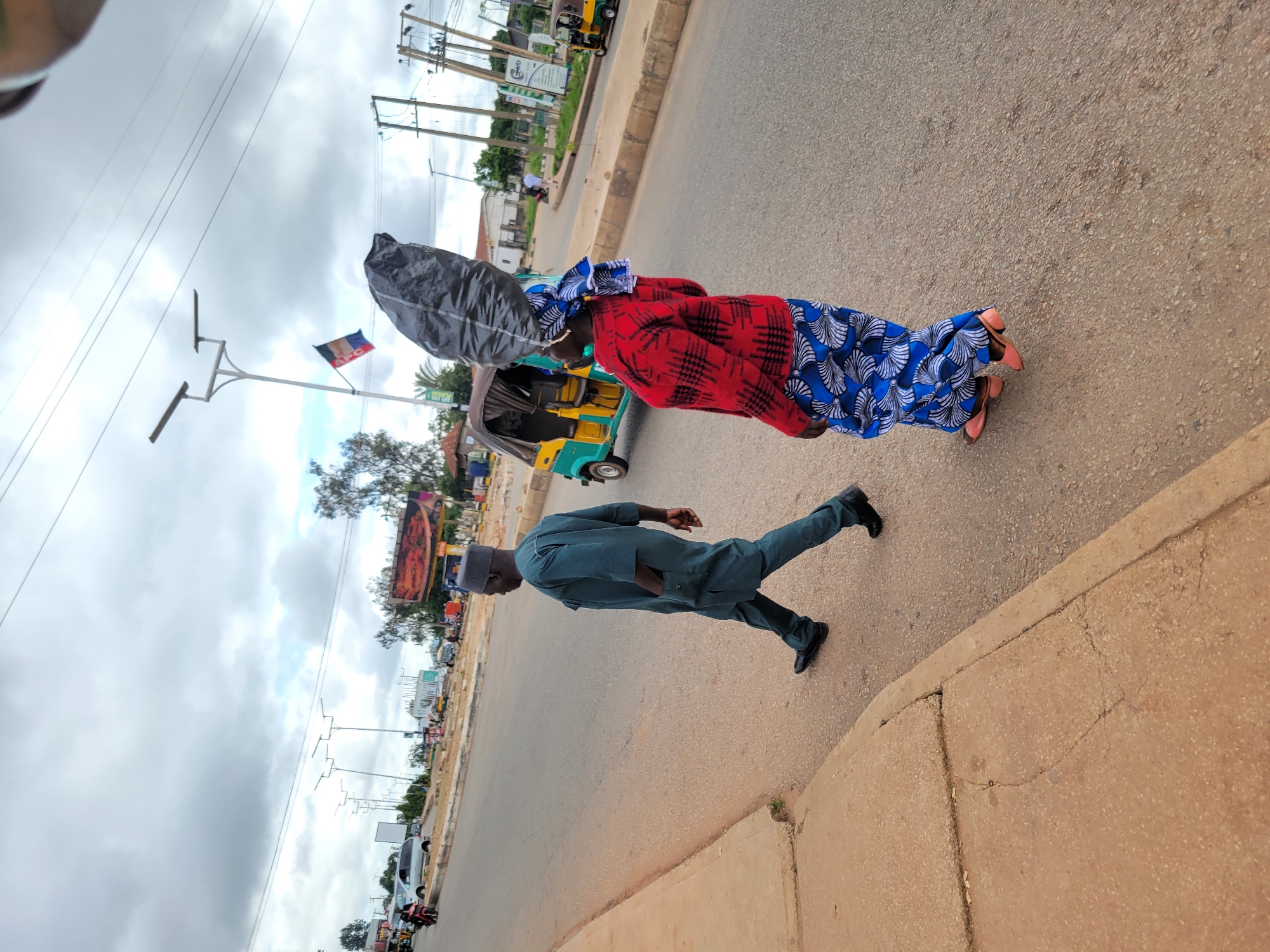 In Jos, Nigeria, a man in a teal traditional outfit walks on the right side of a paved road, his hands in his pockets. To his right, a woman carries a large, black plastic bag on her head, wearing a red and black checkered top and a blue and white patterned skirt. A yellow and green auto-rickshaw is parked behind her, and further down the road, another similar vehicle is visible. The sky is overcast with a mix of gray and white clouds. A flag with red, white, and blue markings is attached to a lamppost in the background. Power lines and streetlights are also present. The roadside features a concrete curb and a brown dirt embankment. Several cars and motorcycles are seen in the distance.