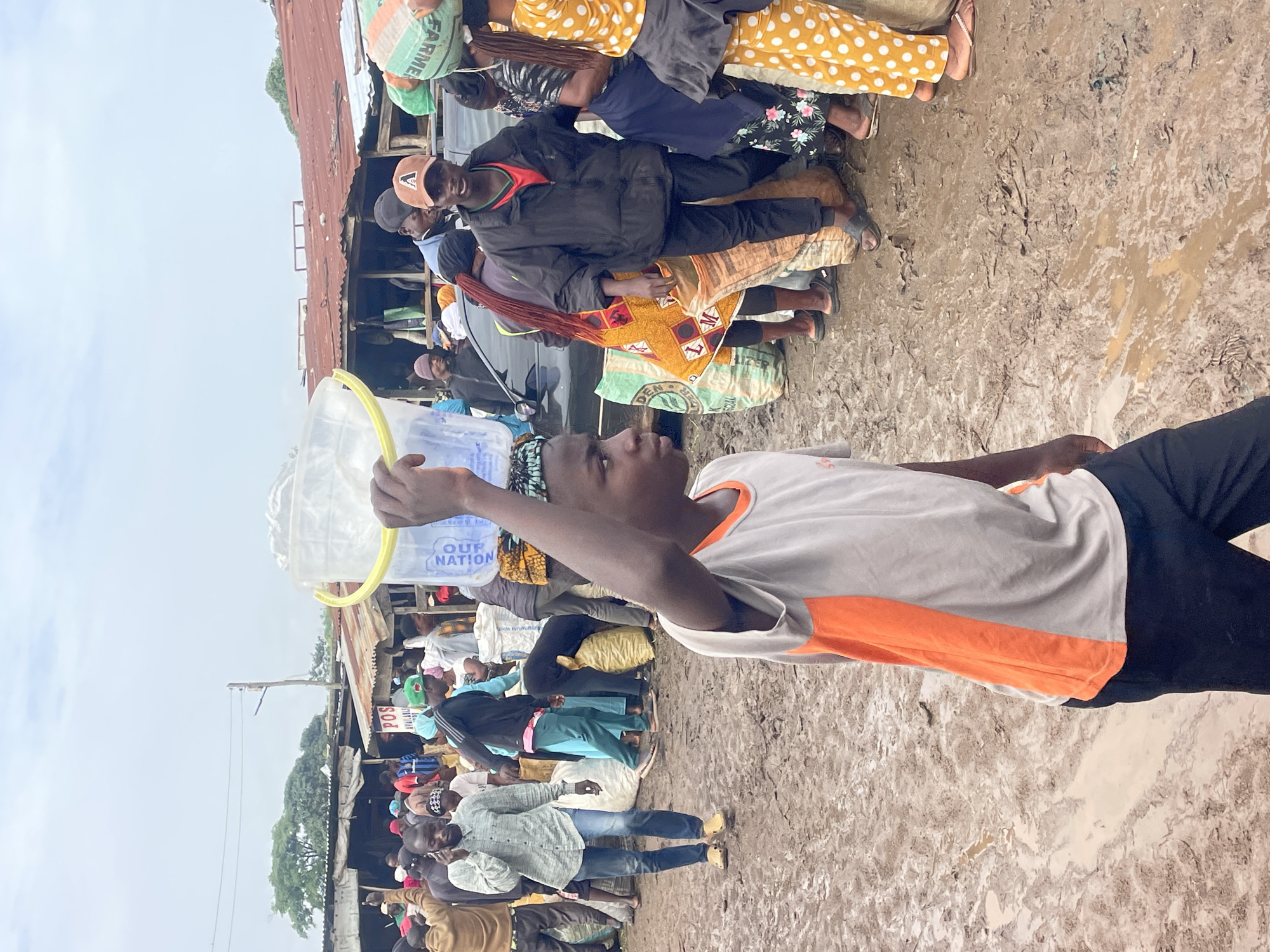 The image depicts a busy outdoor market scene in Gura Top, Nigeria, characterized by muddy terrain and makeshift stalls. A young person in the foreground, wearing a grey and orange t-shirt and dark pants, carries a large clear plastic bucket filled with water on their head. The bucket has a yellow rim and is partially covered. Around them, numerous people are engaged in various activities. To the left, a man in a light blue, long-sleeved collared shirt and jeans is talking on a cell phone, wearing yellow clogs. Behind him, several individuals are visible near stalls made of corrugated metal and wood, some carrying bags.
In the mid-ground and right, the crowd continues. A man in a black puffer jacket and a red t-shirt, wearing an orange baseball cap with a white "A" logo, is looking towards the camera. He stands amidst large sacks. Other individuals are seen in the background, some carrying goods. The ground is heavily saturated with mud, with puddles of water scattered throughout, suggesting recent rainfall or poor drainage. The sky is overcast. The overall impression is one of a vibrant, bustling marketplace despite the challenging environmental conditions.