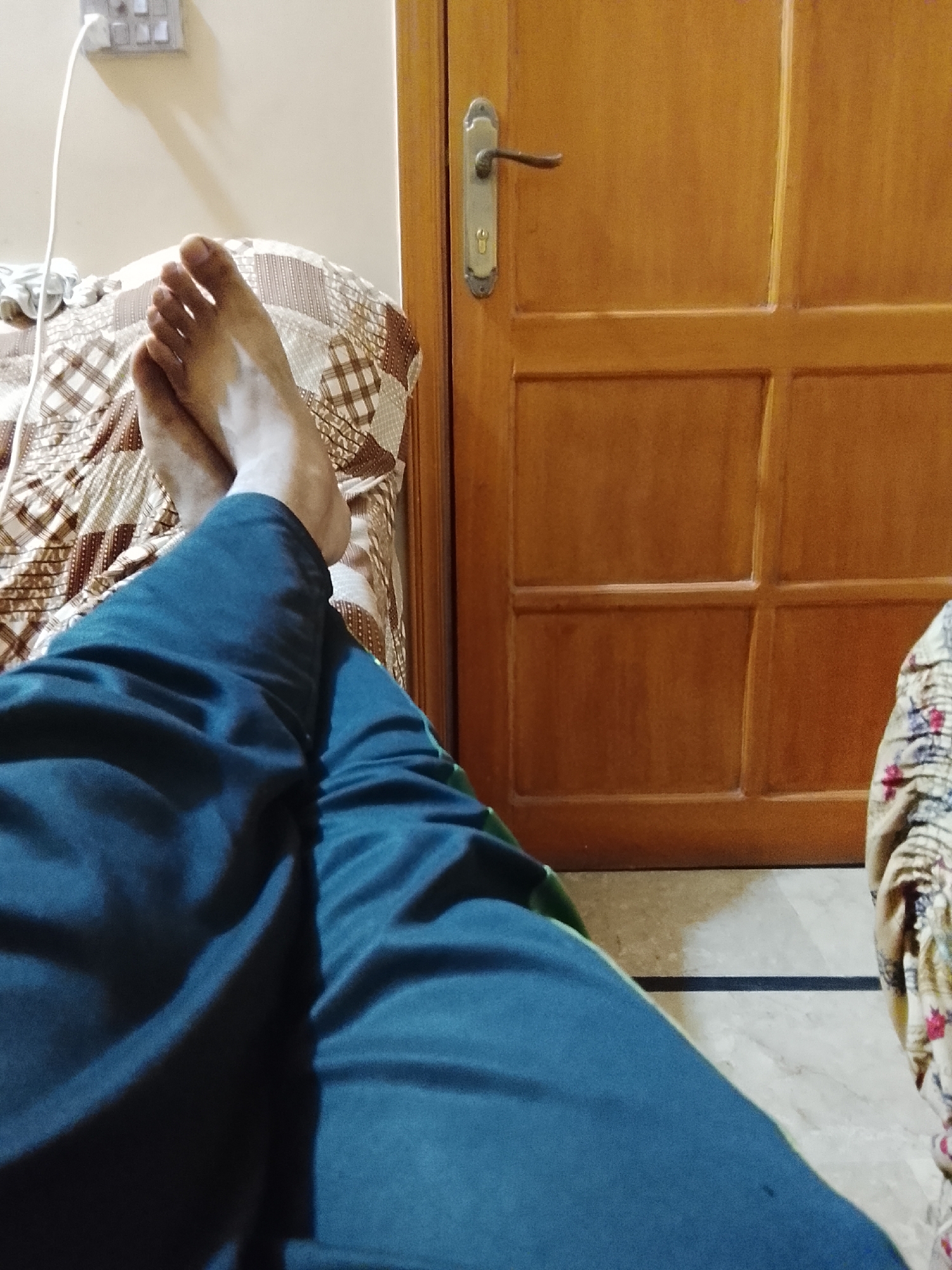 A person is lying on a bed in a room. The person is wearing blue pants. Their bare feet are crossed, with the left foot resting on top of the right foot. The bed has a brown and white checkered patterned bedsheet. A wooden door is in the background, with a metal door handle and lock mechanism visible. A light switch is mounted on the wall, along with a cord plugged into the socket. The scene is within a building in the city of Multan, Pakistan. The flooring visible is a light colored tile.
