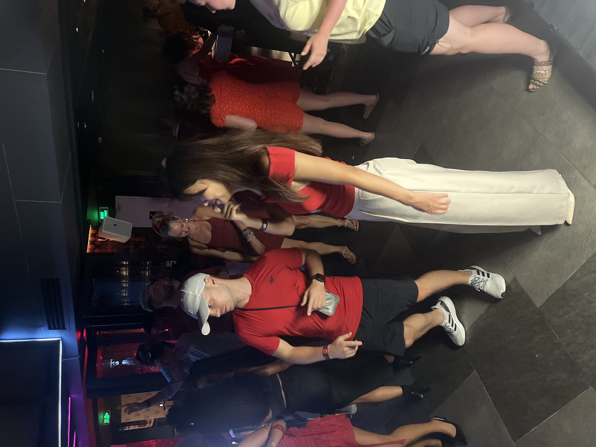 The image depicts a group of people dancing and socializing in a dimly lit nightclub. The location is identified as Đà Nẵng, Vietnam. In the foreground, a man wearing a red t-shirt and black shorts, a white baseball cap, and athletic shoes is looking to his left and gesturing with his right hand. He is holding a gray mask in his left hand. To his left, a woman with long dark hair, wearing a red off-the-shoulder top and white wide-leg pants, is looking towards the left. Behind them, other individuals are visible in various states of dancing and conversation, with some wearing red dresses. The environment features dark walls, a tiled floor, and neon lighting accents along the ceiling, creating a typical nightclub ambiance. An exit sign with green lettering is visible in the background.