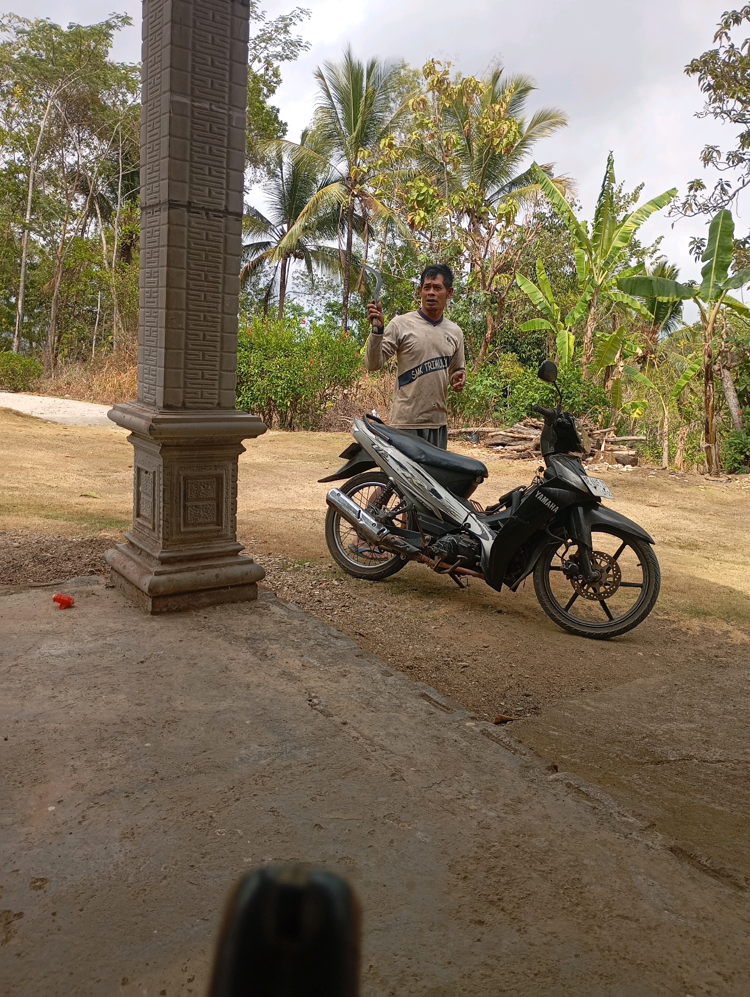 A middle-aged Indonesian man stands next to a black and silver motorcycle. He is wearing a light brown long-sleeved shirt with "SMK TRIHULULI" printed on the chest, and dark shorts. He is holding a sickle in his right hand and appears to be speaking. The motorcycle is parked on a dirt and grass area in front of a concrete structure that resembles a pillar or a small building. The pillar is ornate with carved patterns. Behind the man and motorcycle, there are numerous trees, including palm trees and banana trees, and a cloudy sky. The scene appears to be outdoors in a rural or semi-rural area. The location is specified as the city of Ketepung, Indonesia.