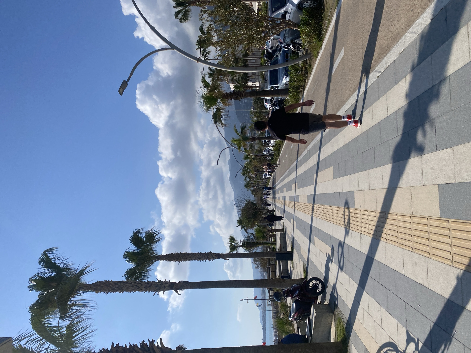 The image shows a vibrant, sunny scene on a coastal promenade in Alanya, Turkey. The view is looking down a wide, tiled walkway, flanked by palm trees and streetlights.
In the foreground, a man walks with his arms slightly outstretched, wearing a dark t-shirt, shorts, white socks with red stripes, and red sneakers. His long shadow, along with shadows from the streetlights, stretches across the patterned grey and beige paving, indicating bright midday sun. A distinct yellow tactile paving strip runs along the center of the promenade.
To the right, a row of tall palm trees and modern streetlights line the path, with parked cars and other vehicles visible behind them. Further in the background, majestic mountains rise, partially obscured by more palm trees and buildings. The sky is a clear, brilliant blue with large, fluffy white clouds.
To the left of the promenade, glimpses of a blue sea or bay are visible, confirming the coastal setting. Several other people are visible walking along the promenade in the distance, enjoying the pleasant weather. A motorcycle is parked beside the walkway in the middle ground. The scene conveys a leisurely atmosphere in a scenic, warm environment. There is no visible text in the image.