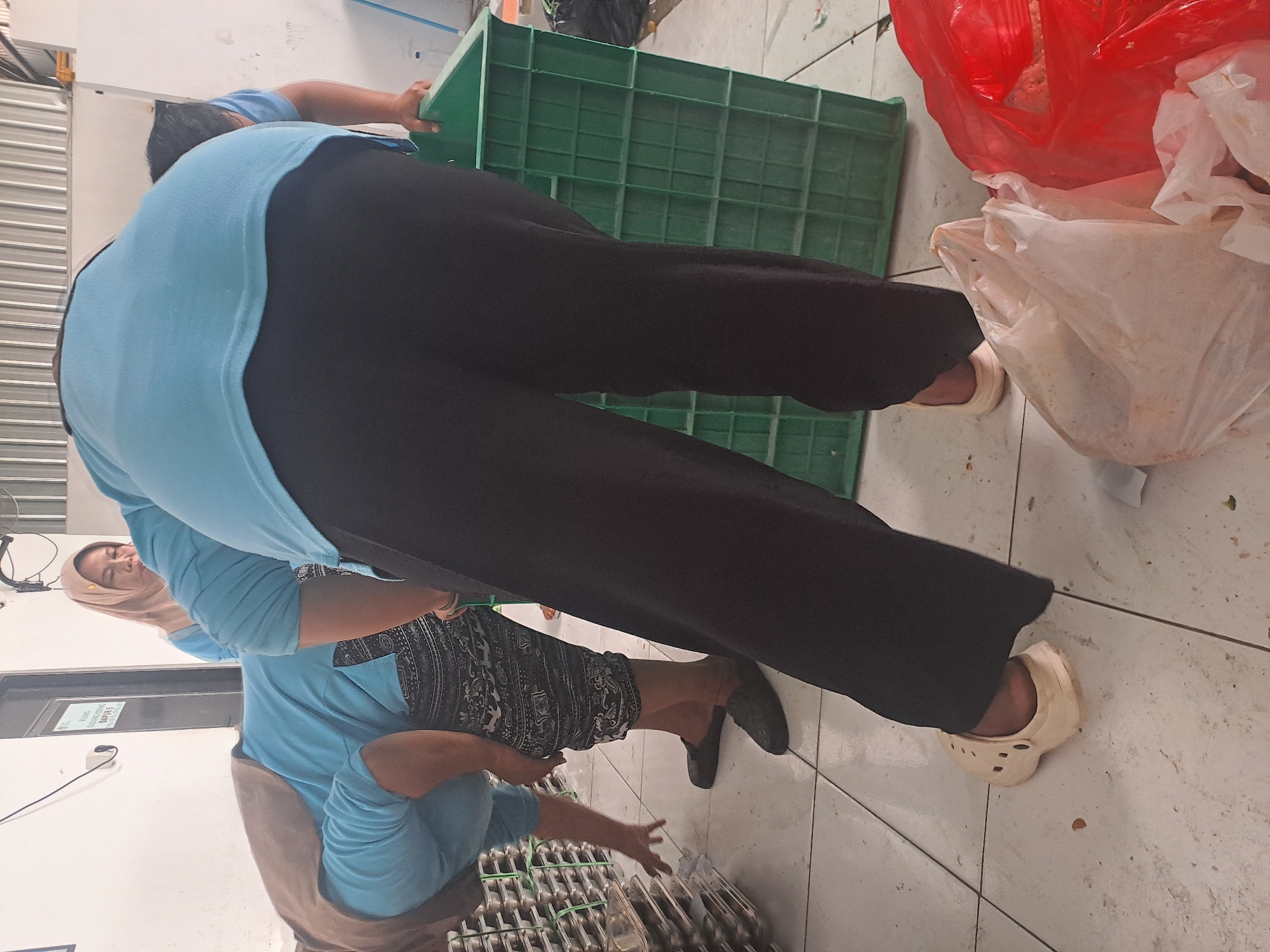 In Mojorejo, Indonesia, three individuals are engaged in an activity within a tiled room, likely a food preparation or processing area. The person in the foreground, wearing black pants and light-colored Crocs, is bent over, partially obscuring a large green plastic crate. Behind them, another person in a blue shirt and patterned pants is also bent over, reaching towards stacks of metal trays. A third individual, wearing a light-colored headscarf and blue shirt, stands to the left, observing. The environment appears functional, with a corrugated metal ceiling visible in the background and various items, including plastic bags and discarded materials, on the floor. The overall impression is one of work being done, possibly related to food handling or packaging, in a somewhat cluttered industrial or commercial setting.