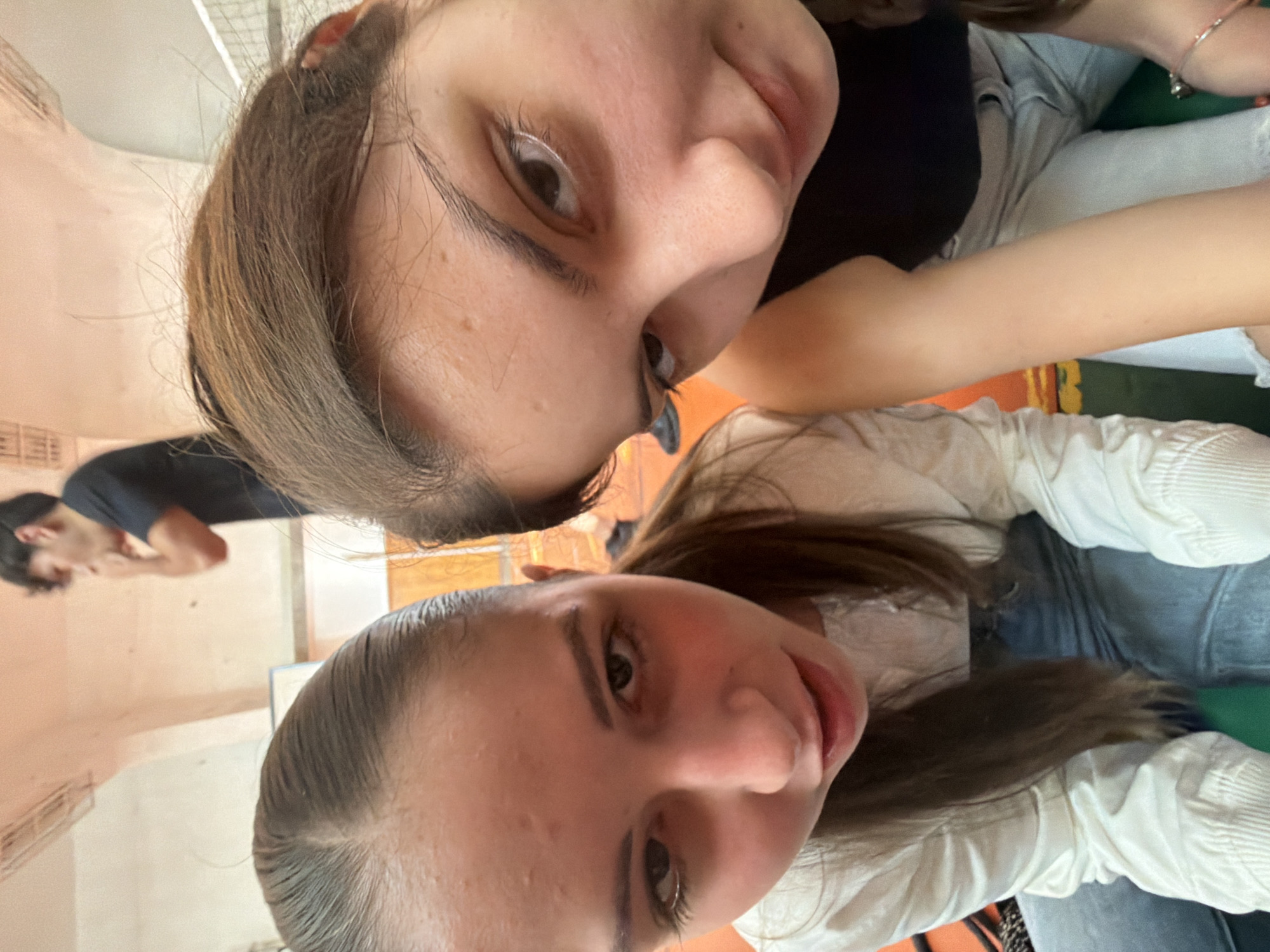An indoor, upward-angle selfie features two young women in the foreground, with a young man visible in the background. The image is oriented vertically, with the women positioned to appear side-by-side or stacked from the camera's perspective.
The woman closer to the top of the frame has long, light brown hair, fair skin, and is wearing a dark, possibly black, top. She is looking towards the camera with a soft expression, her right eye prominent. A faint bracelet is visible on her wrist. The second woman, closer to the bottom, has dark brown hair pulled back from her face and fair skin. She is wearing a light-colored, long-sleeved, ribbed top and light blue jeans. She also looks towards the camera with a relaxed expression, revealing a small earring.
In the background, out of focus, a young man with dark hair and a dark t-shirt is partially visible, appearing to be looking down or away from the camera. The setting is a casual indoor space, possibly a room with light peach-colored walls. Elements of what appear to be furniture or seating, including a green cushion and an orange surface, are visible around the women. The overall mood suggests a friendly and relaxed interaction, possibly among friends. There is no visible text in the image. The location context provided is Burgas, Bulgaria.