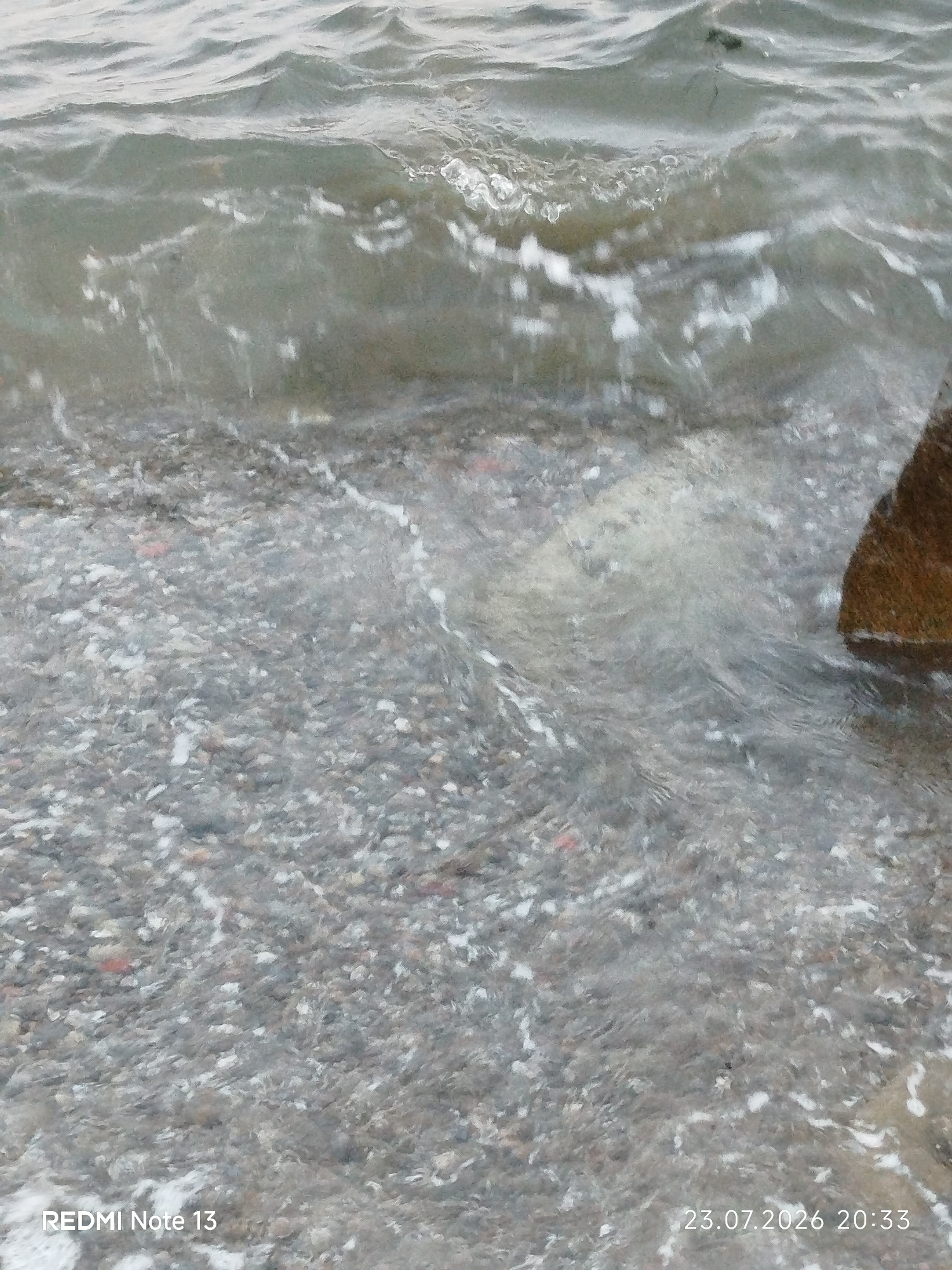 The image shows a close-up view of shallow, choppy water lapping over a pebble-strewn shore. The water is murky and reflects the overcast sky. White foam from the waves is visible against the dark pebbles. In the upper right corner, a portion of a dark, reddish-brown rock is present. The bottom left corner displays the text "REDMI Note 13" and the bottom right corner displays "23.07.2026 20:33". The scene appears to be a natural shoreline, likely a beach or the edge of a body of water, possibly within the Sithonia Municipal Unit, Greece, given the date stamp format which could correspond to a location in Europe. No people or specific landmarks are visible to pinpoint the exact location within Sithonia.