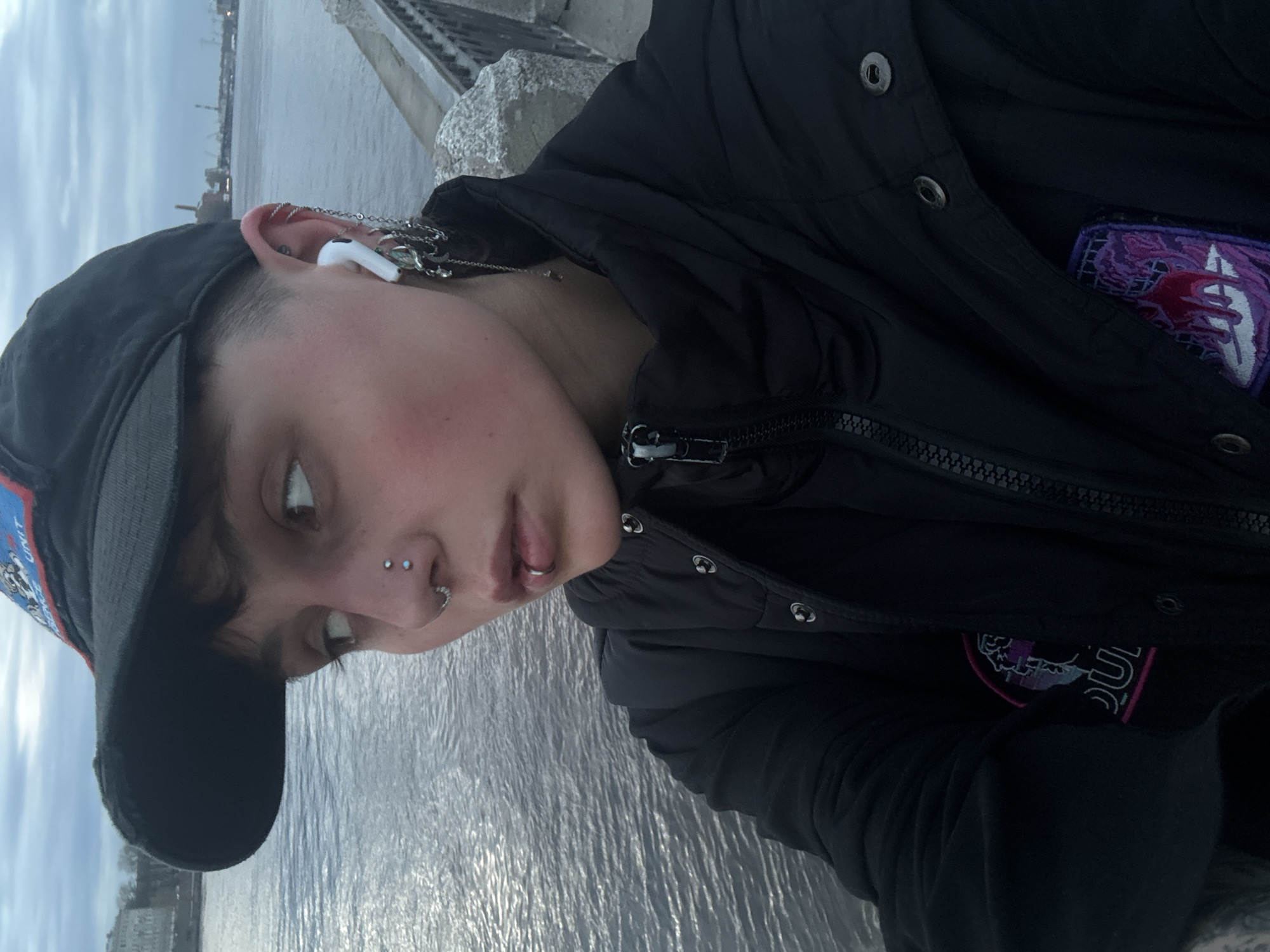 A close-up, slightly angled portrait captures an individual outdoors, set against a backdrop of a large body of water. The person has short dark hair, a fair complexion, and is wearing a dark baseball cap with a colorful patch on its side. They are dressed in a dark, possibly quilted or puffy jacket, which features a prominent zipper and snap buttons.
The individual showcases multiple facial and ear piercings. On their nose, two studs are visible near the bridge/septum area, along with a ring on the nostril. A silver ring adorns their lower lip. Their ear is heavily decorated with various piercings, including studs and rings, some interconnected by delicate chains. A white earbud is inserted into one ear. A vibrant, embroidered patch with purple and pink tones and a stylized, partially visible logo (featuring shapes and possibly a 'U') is on the jacket's chest.
The background shows a wide body of water, likely a river or canal, its surface rippled under an overcast sky. A concrete or stone embankment with a railing or fence runs along the water's edge. In the far distance, faint outlines of buildings and what appear to be industrial structures or cranes are visible beneath the cloudy conditions. The person's expression is neutral as they look slightly away from the camera. The scene depicts an urban waterfront in Saint Petersburg, Russia, under cloudy skies.