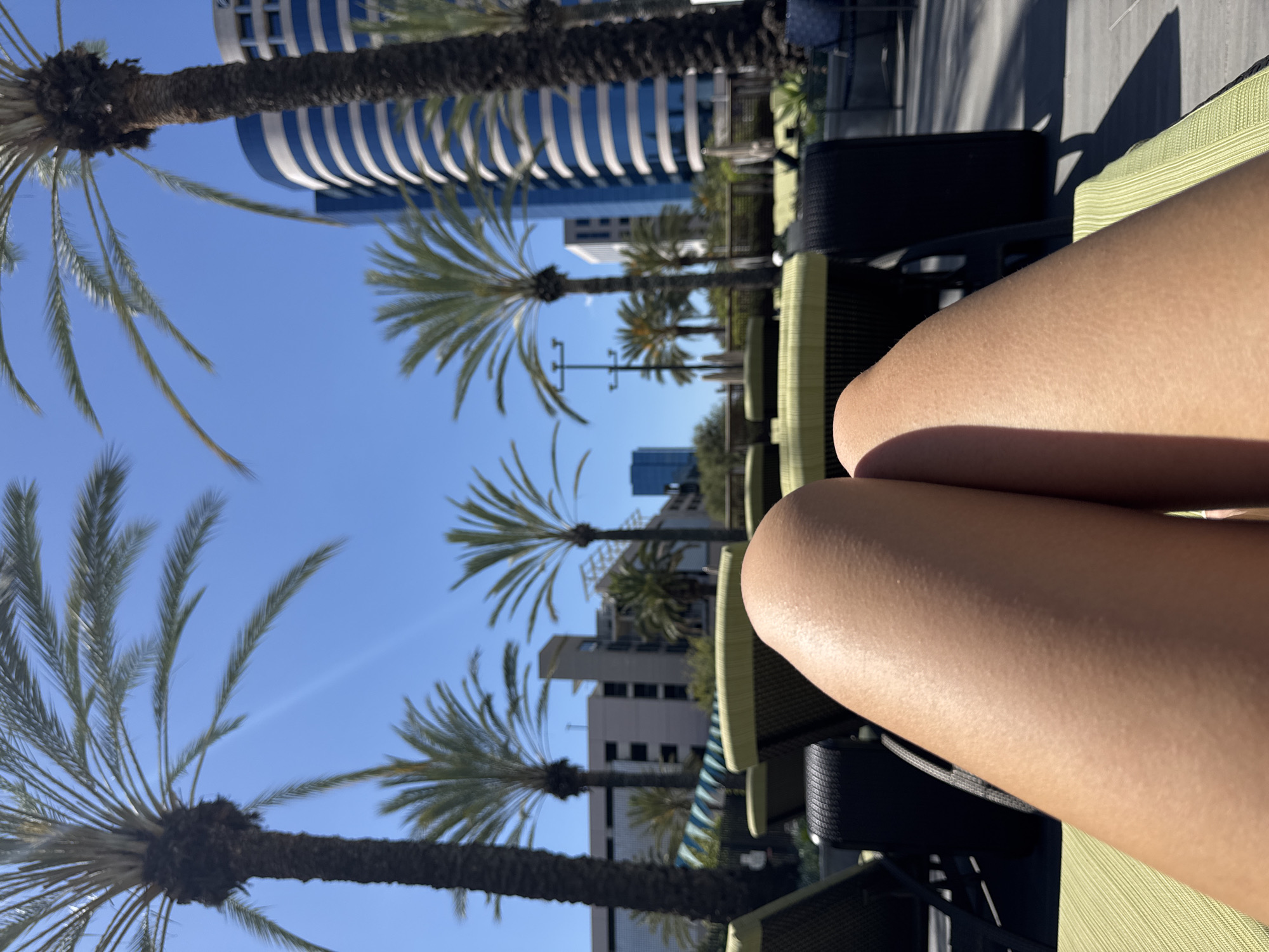 The image shows a first-person perspective of someone sitting on a lounge chair, presumably outdoors. The person's bare legs are in the foreground. The background features palm trees against a clear blue sky, with various buildings visible in the distance. The location is strongly suggested to be in Irvine, United States of America, due to the presence of palm trees and modern buildings, which are typical of the area. The lounge chair appears to be made of a woven material with a light green color. The buildings in the background vary in height and architectural style, with one particularly prominent building displaying a curved facade with blue and white horizontal stripes.