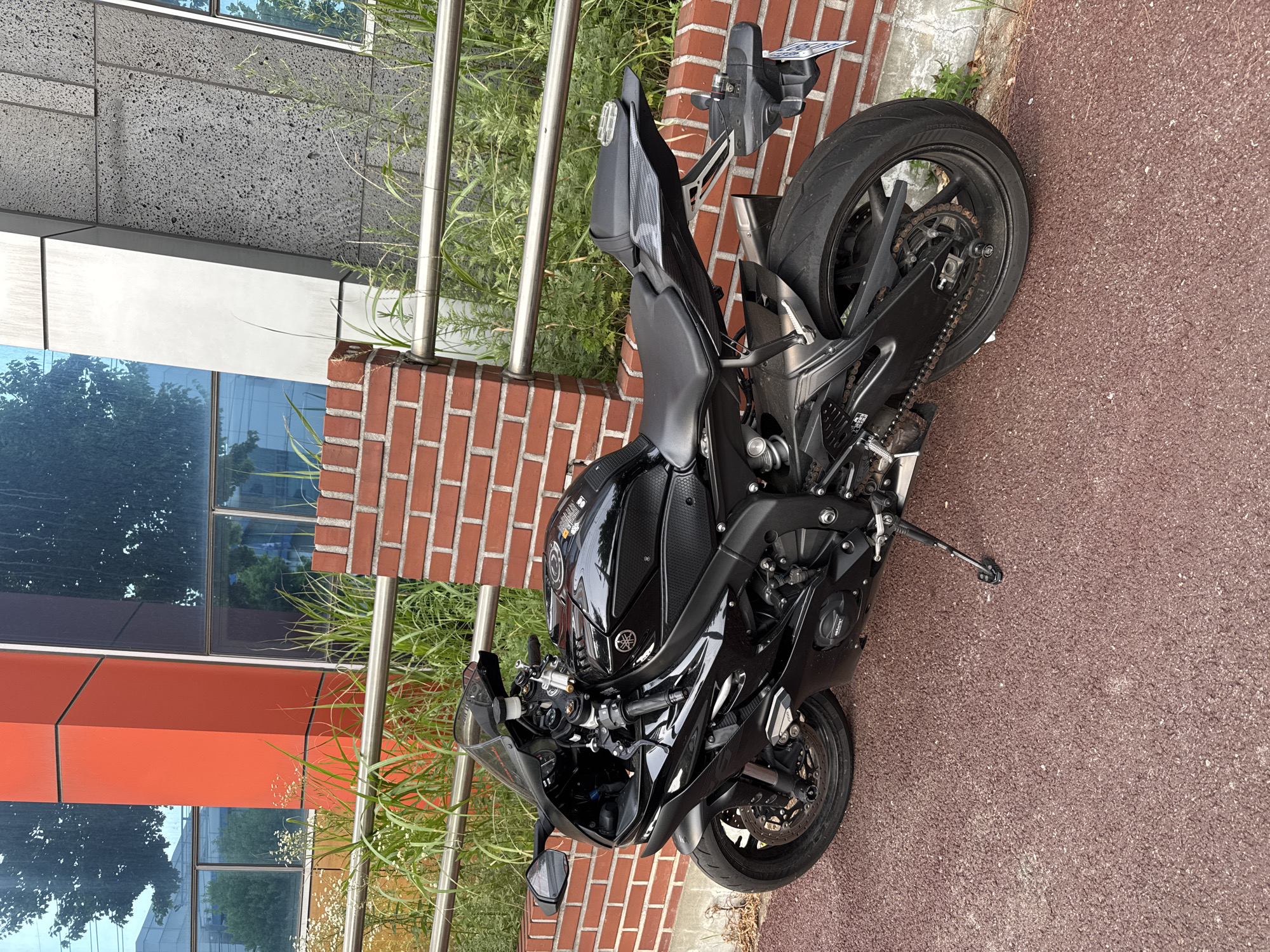 A black Yamaha motorcycle is parked on a reddish-brown paved surface in front of a building with red brick and dark gray textured walls, and large blue-tinted windows. The motorcycle is positioned at an angle, facing left, with its kickstand down. The building's facade features an orange vertical beam on the left side. A metal railing runs horizontally in front of the building, with tall green grass and some white wildflowers growing behind it. The scene appears to be outdoors, in a moderately urban setting, possibly a commercial or institutional area in Pyeongtaek-si, South Korea, given the context provided. No people are visible in the image.