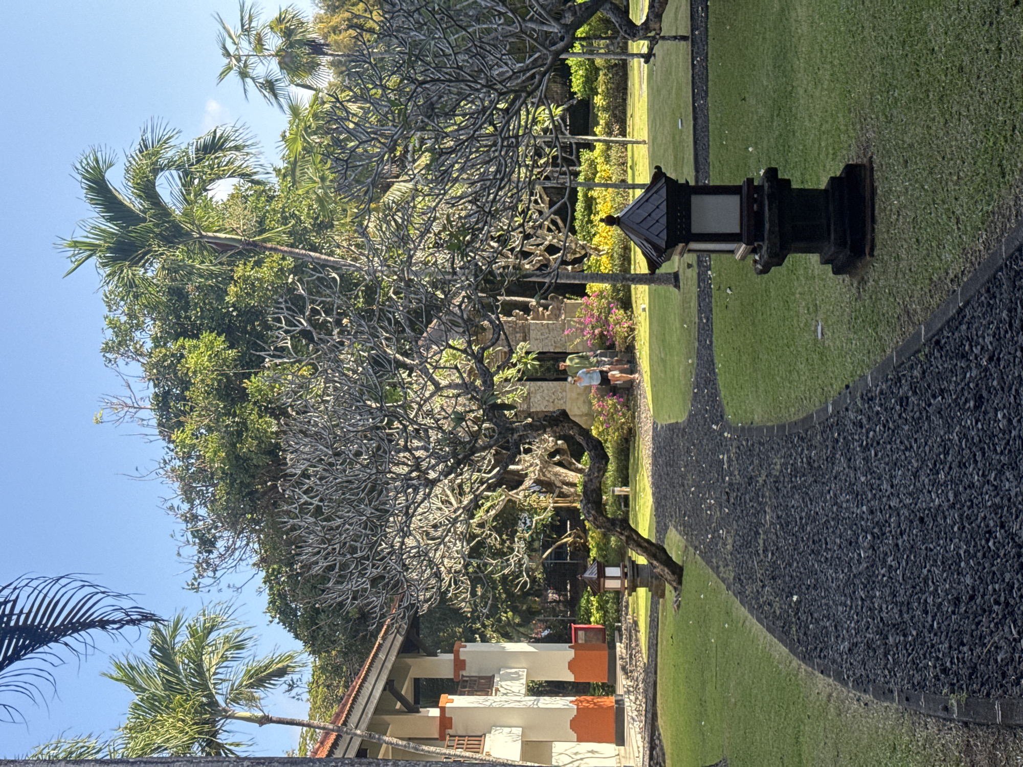 The image depicts a paved pathway made of dark gravel curving through a manicured lawn. On the left, a building with white and orange accents and balconies is visible. In the background, lush green trees and palm trees dominate the landscape, under a clear blue sky. Two individuals, one wearing a green shirt and shorts, and the other a blue top and shorts, are walking on the pathway in the middle ground. Several traditional-style lanterns are placed on the lawn. The scene appears to be a resort or hotel garden in Kuta Selatan, Indonesia, characterized by tropical vegetation and architecture.