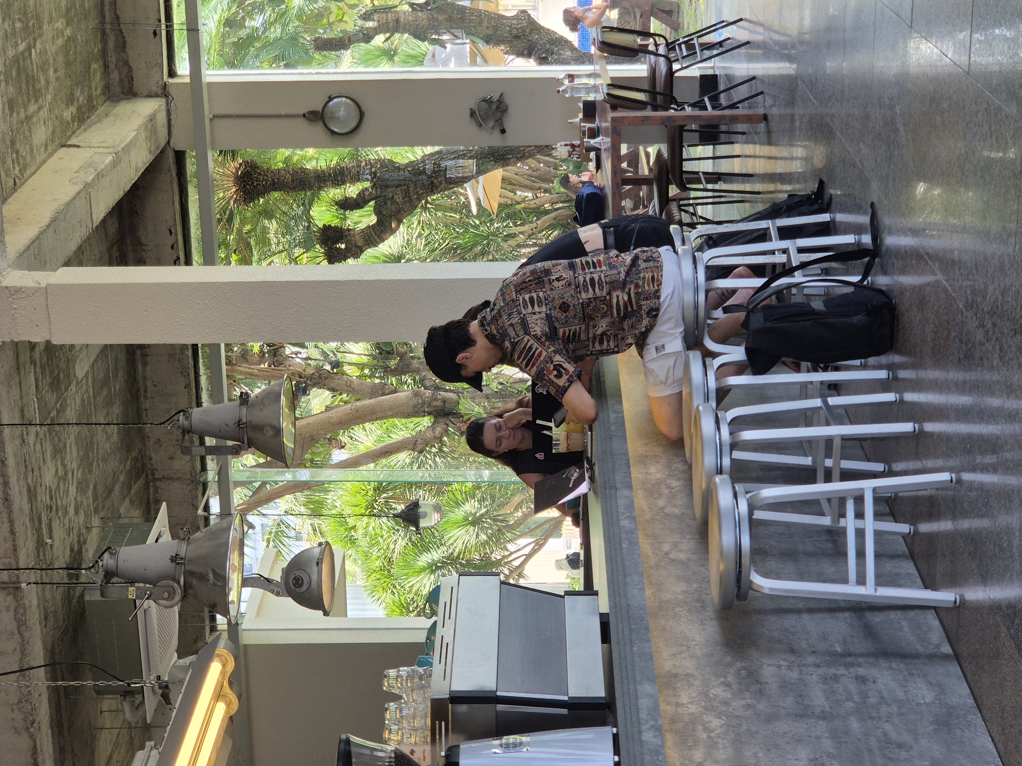 The image captures a scene inside a modern cafe, likely located in the city of Da Nang, Vietnam, characterized by its open-air design and lush tropical surroundings visible through large glass windows. In the foreground, three white bar stools with silver seats are arranged along a grey, concrete-like counter. A black backpack rests on the floor next to the closest stool.
Behind the counter, a woman with long dark hair is seated, facing a laptop, with her hand resting on her chin, appearing contemplative. To her right, a man wearing a black baseball cap and a colorful, patterned short-sleeved shirt is also seated at the counter, looking down at his phone. His white shorts and the detail of his patterned shirt are prominent.
In the background, beyond the counter and through the large windows, a vibrant green tropical landscape with palm trees is visible, suggesting an outdoor seating area or courtyard. A few tables and chairs are set up outside. Other patrons can be seen faintly in the distance, including a woman sitting at a table outdoors. The interior of the cafe features exposed concrete ceilings with industrial-style pendant lights, and pillars supporting the structure, contributing to a contemporary, minimalist aesthetic. The overall atmosphere seems relaxed and casual, with people engaged in personal activities or social interactions.