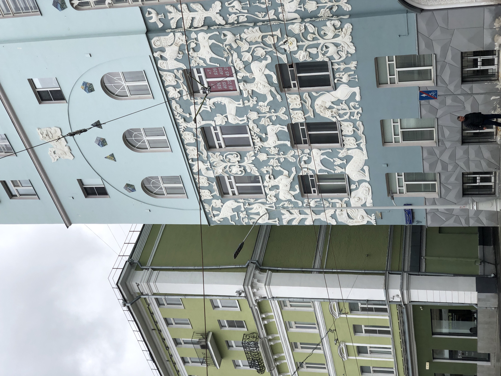 The image shows two buildings in Moscow, Russia. The top building is light blue and gray with white decorative carvings of animals and plants. The bottom building is green with white trim and a black wrought-iron balcony. Electrical lines run between the buildings. A person is standing on the sidewalk in front of the light blue and gray building.