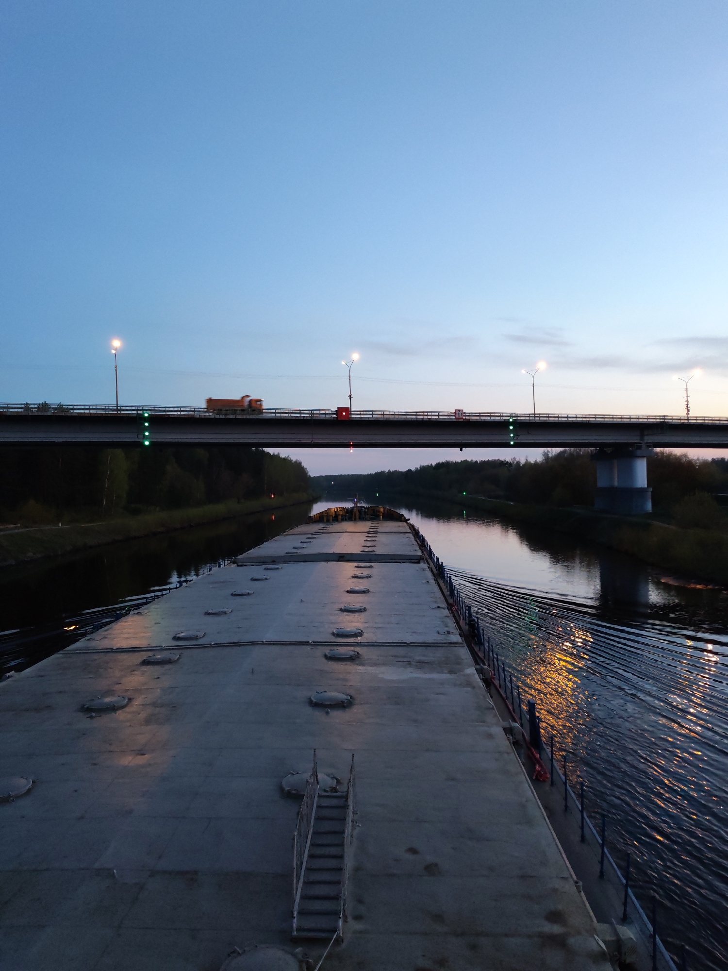 The image depicts a view from the deck of a large barge or boat moving along a canal under a bridge at dusk. The barge deck is gray and relatively featureless except for evenly spaced round hatches and a set of stairs. The canal water is dark and reflects light from the opposite bank. The bridge has three lit street lamps and a truck is visible on the bridge. The location is in the city of Dmitrov, Russia. The environment consists of the canal, trees lining the banks, and the evening sky.
