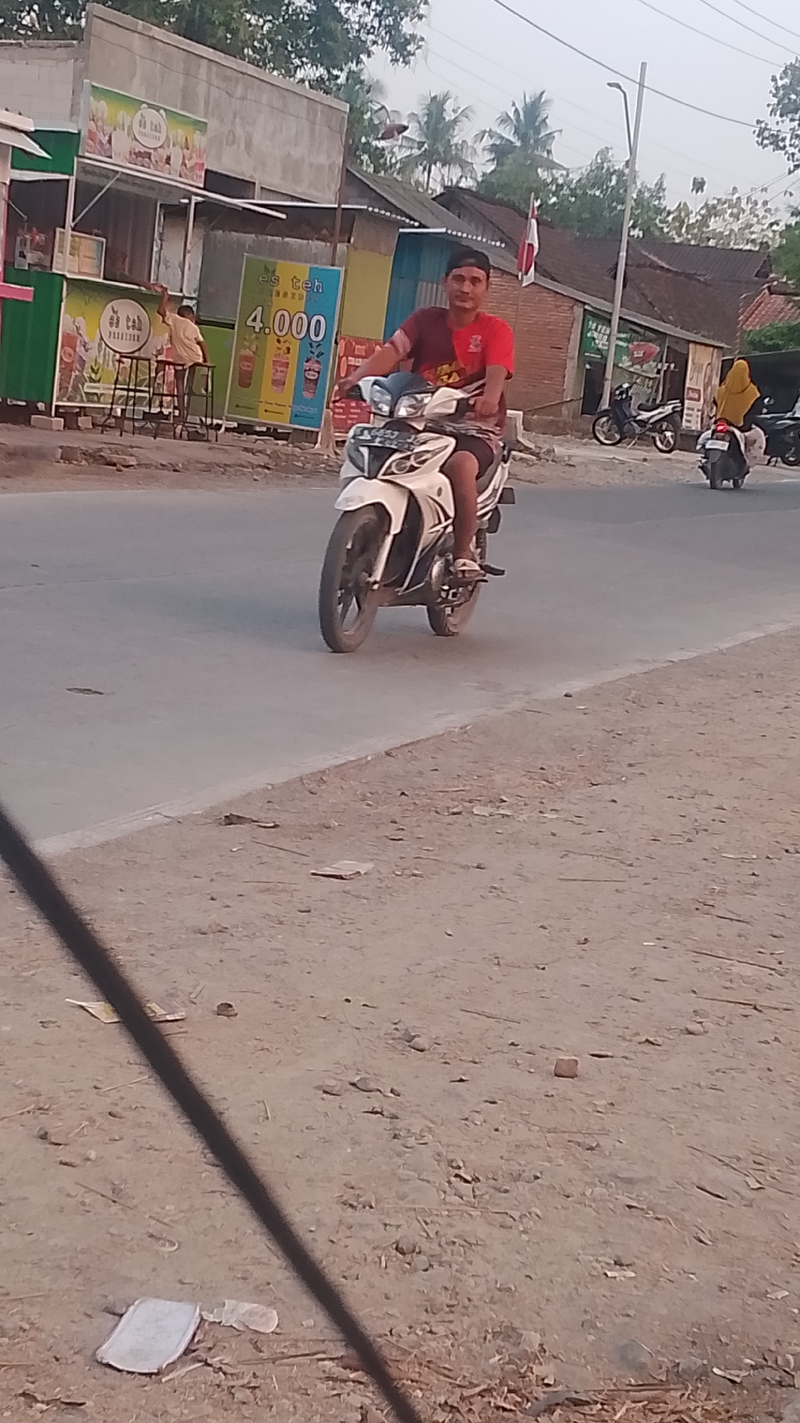 In Karanglangu, Indonesia, a person is riding a white and black motorcycle on a paved road. The rider is wearing a red and black t-shirt and dark shorts. In the background, there are several small buildings and stalls. One stall has a sign advertising "es teh" for 4,000. Another person, wearing a yellow headscarf, is riding a black motorcycle in the distance, along with another person on a motorcycle behind them. The scene appears to be a street in a village or small town, with a mix of residential and commercial structures. The overall lighting suggests it is daytime, possibly late afternoon.