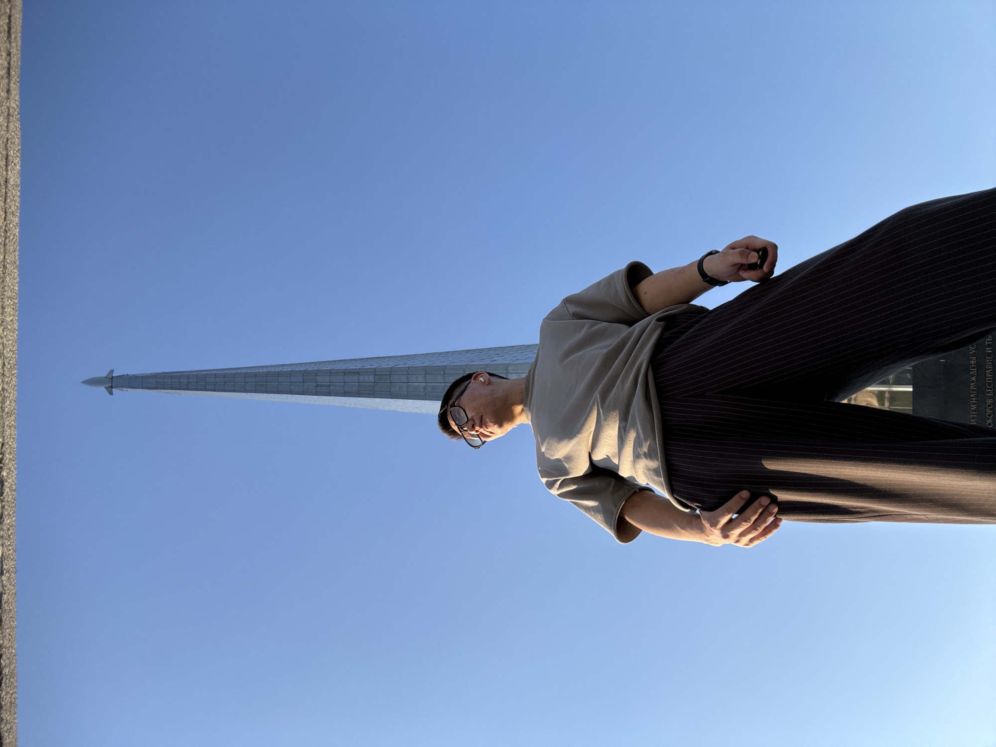 The image shows a young man standing outdoors, looking up at a tall, slender monument topped with a rocket. The monument appears to be made of metal panels and tapers to a sharp point against a clear blue sky. The man is wearing glasses, a grey t-shirt, and dark pants with vertical stripes. He has his left hand resting on his hip and his right hand is slightly raised. The low angle of the shot emphasizes the height of the monument and the upward gaze of the man. Some text is visible on the base of the monument in the lower right portion of the image, but it is illegible. The setting is likely a public space in Moscow, Russia, given the context of space-related monuments which are common in the city.