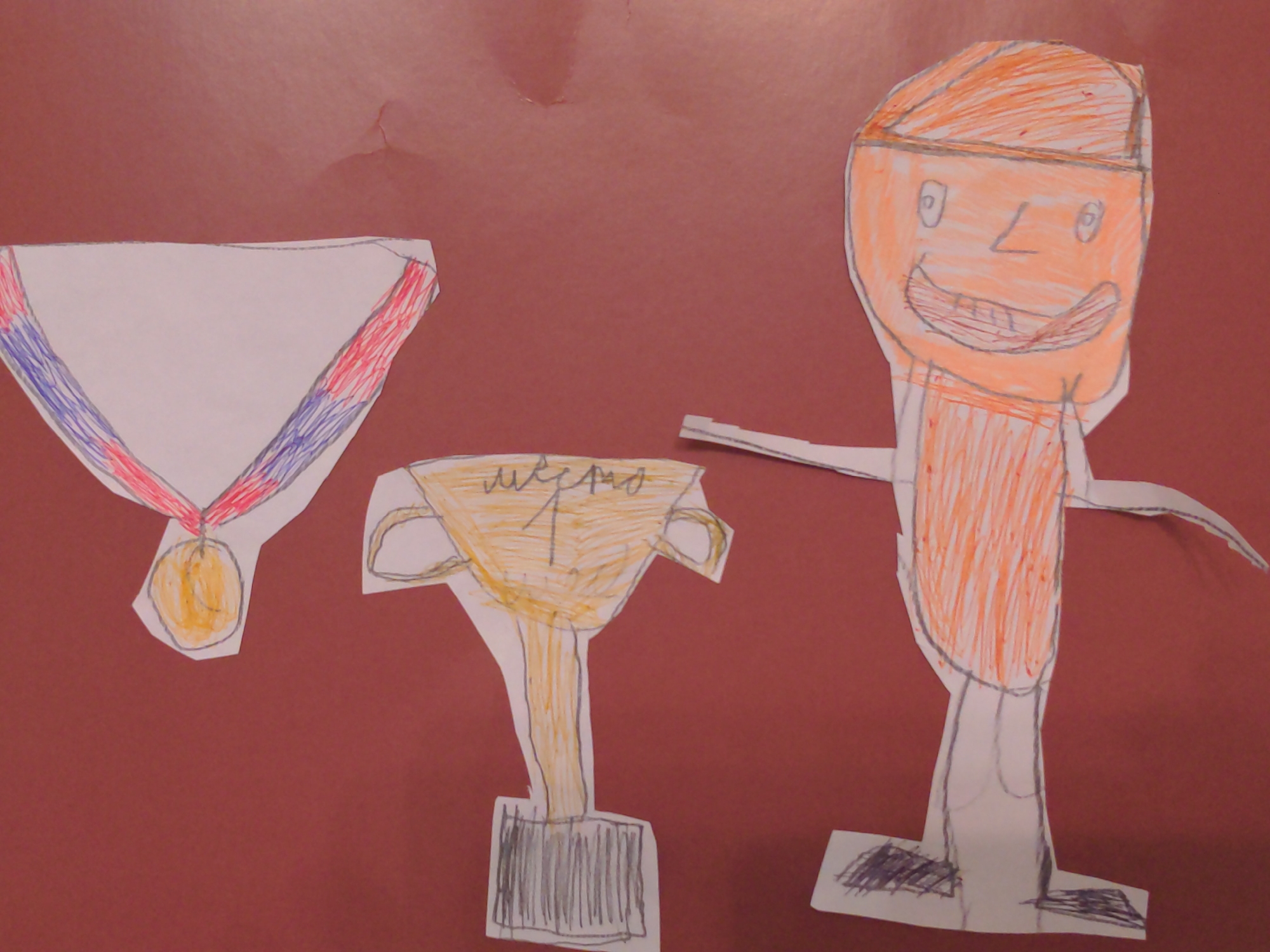 On a reddish-brown background, a child's drawings have been cut out and arranged. From left to right, there's a medal with a red and blue striped ribbon attached to a gold medal, a gold trophy with "1 место" written on it, and an orange figure with a smiling face. The drawing appears to be laid out on a flat surface. There is no indication of the city of Moscow, Russia.