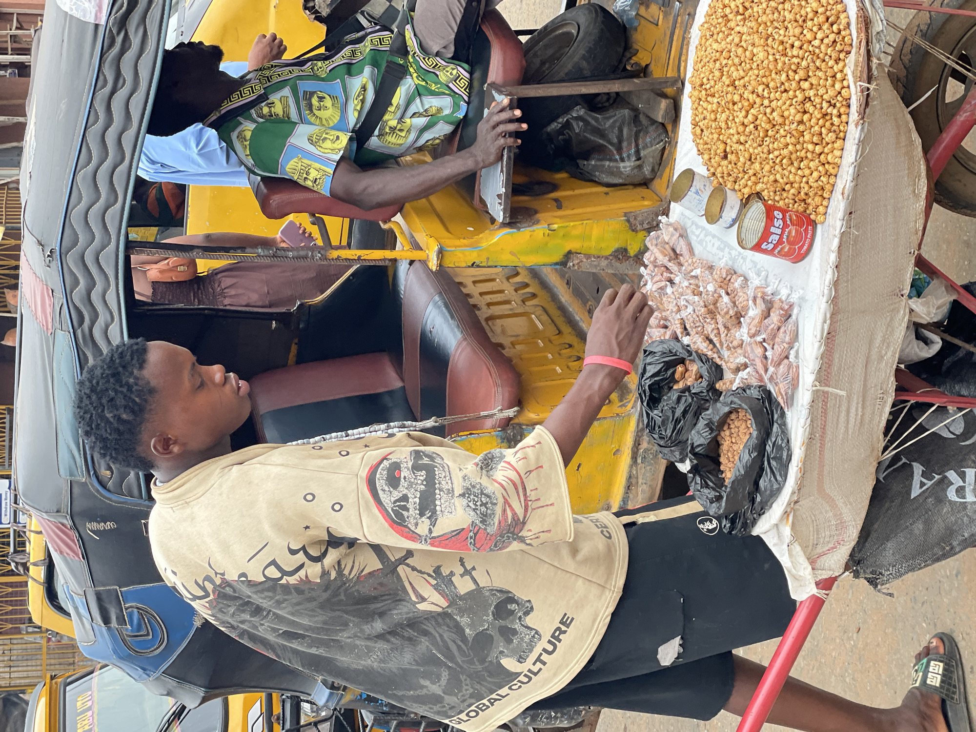 In Nkpor, Nigeria, a yellow tricycle is parked along a street, with two men visible in and around it. One man, wearing a tan sweatshirt with skull designs, is standing next to the tricycle, his arm extended towards a display of peanuts and other items for sale. Another man is seated inside the vehicle, wearing a green and yellow patterned shirt with images of Egyptian pharaohs. He is holding a panel or lid in his hand. The tricycle serves as a mobile vending unit, laden with peanuts, packaged snacks, and canned goods. The vehicle's interior reveals brown leather seats. The surrounding environment consists of urban street elements, including other vehicles.