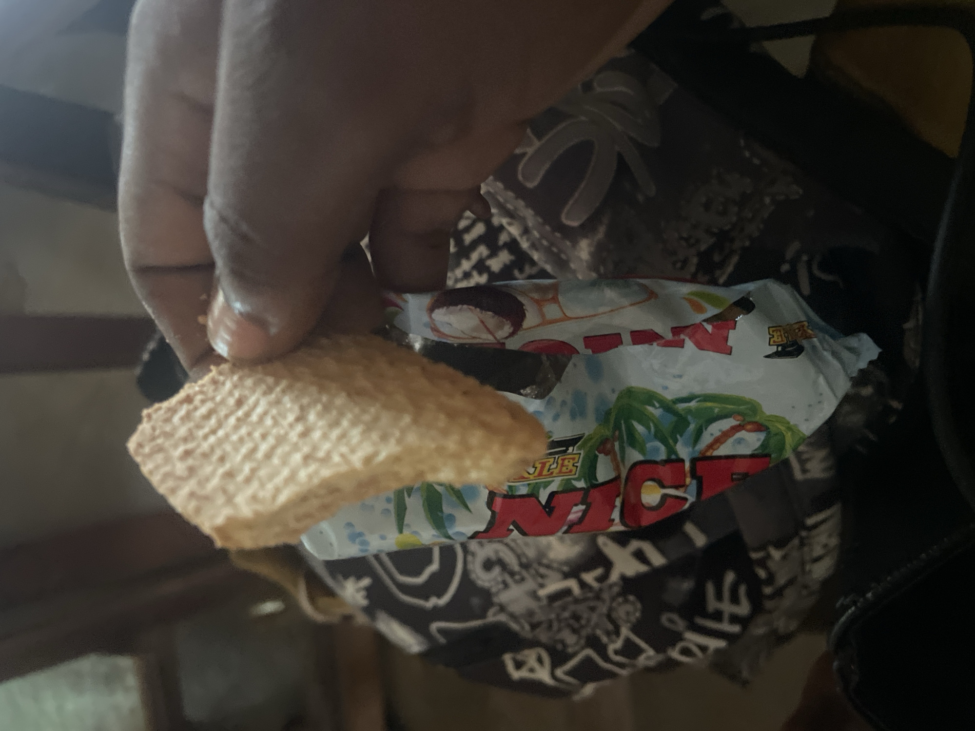A hand, likely belonging to a person of color, is holding a square-shaped, textured snack. The snack appears to be a wafer or cracker with a grid-like pattern. The hand is positioned over a brightly colored wrapper that contains text and imagery. Part of the wrapper displays the word "NICE" in large, red, stylized letters, with a yellow circular element to the left of the letter "C." Behind the "NICE" text, there is a depiction of palm trees and a tropical sky. Below this colorful section, the wrapper has a black and white pattern with various symbols and shapes. The scene is dimly lit, suggesting an indoor setting or an evening time. The overall impression is casual, possibly someone enjoying a snack. The location context provided is Lokoja, Nigeria.
