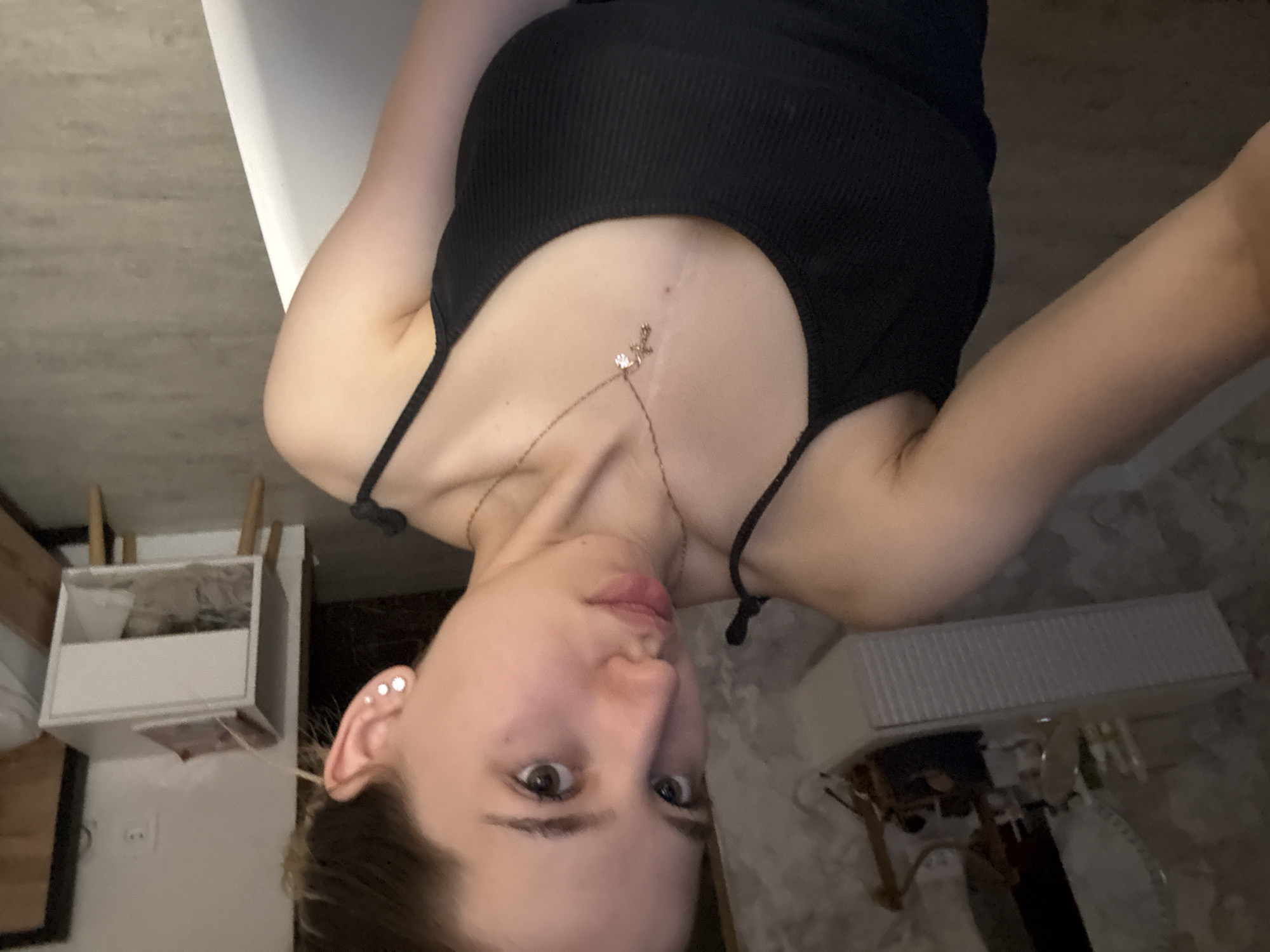 A young woman with fair skin, light brown hair, and multiple piercings in her left ear is posing for a selfie. She is wearing a black tank top and a delicate gold necklace with a small pendant. A thin, light-colored scar is visible on her chest, extending downward from the base of her neck. The background appears to be an interior setting, possibly a bathroom, with white tiled floors and a vanity. A white storage unit with compartments is visible on the left side of the image. No specific information indicates the location is in the city of Noviy, Russia.