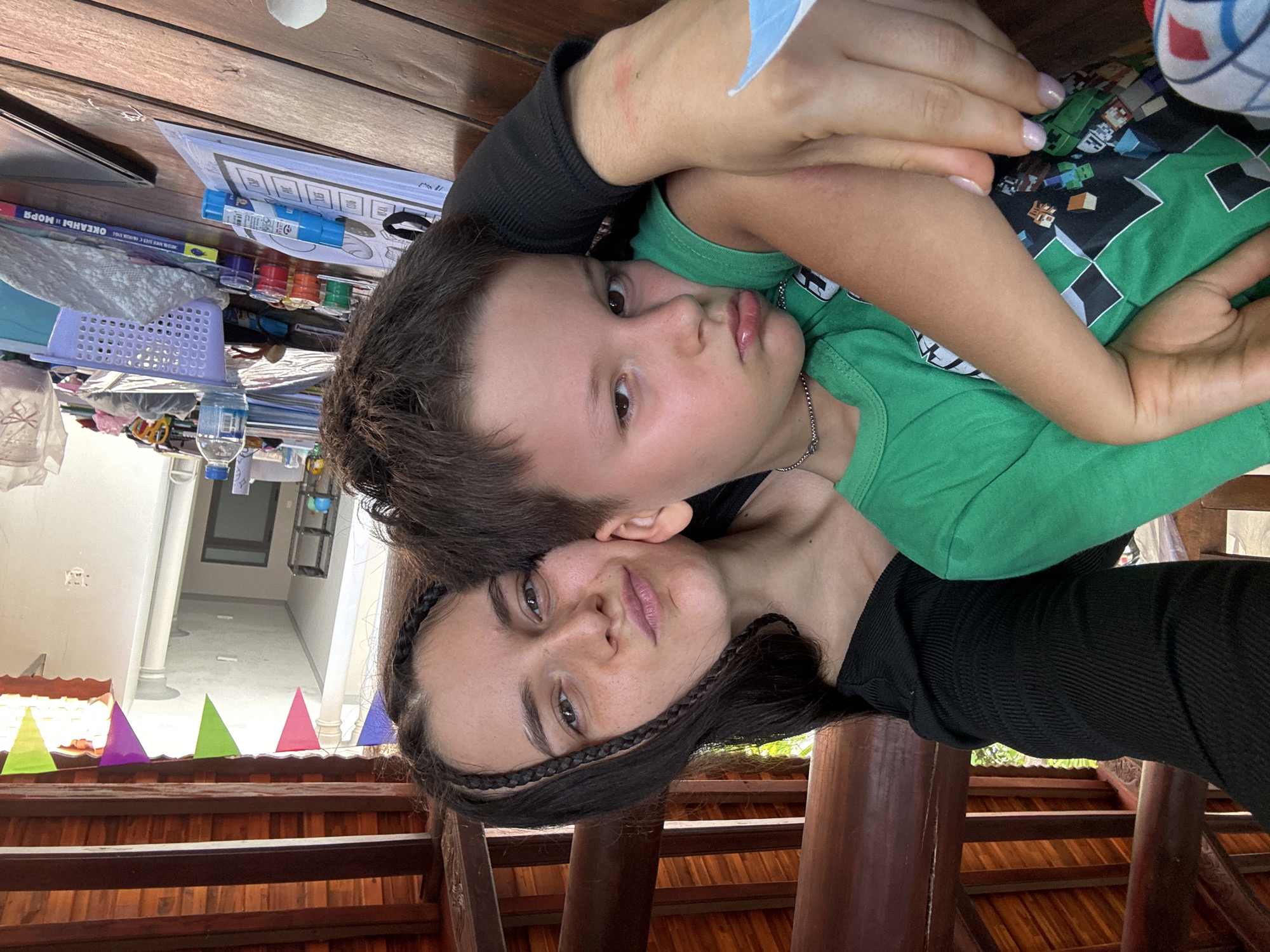 A closely framed portrait captures an adult woman embracing a young boy in what appears to be a covered outdoor space in Mũi Né, Vietnam. The image, originally taken in portrait orientation and rotated, shows the woman's head resting against the boy's, both looking towards the camera.
The woman has long, dark hair, with braided sections framing her face, and a gentle, slightly smiling expression. She is wearing a black ribbed top. Her left arm is wrapped around the boy, and her manicured nails are visible. The young boy, possibly around 5-8 years old, has short dark hair and brown eyes, looking directly at the camera with a neutral to curious expression. He wears a green t-shirt, which appears to have a pixelated, possibly Minecraft-themed, pattern on its shoulder and sleeve. A thin silver chain is visible around his neck.
In the foreground, colorful triangular bunting in shades of green, purple, pink, blue, and yellow is strung across wooden beams, suggesting a festive or playful atmosphere. Behind them, a wooden table is cluttered with various items, including art and craft supplies: small pots of paint (red, orange, green, blue), markers, and paper. A light purple plastic basket and a plastic water bottle are also on the table. A book or notebook with Cyrillic text, reading "OKEAHbl И MOPЯ" (Oceans and Seas), is visible among the items. The background shows a light-colored wall or building, with an open doorway leading to a brighter area. The lighting suggests it is daytime.
