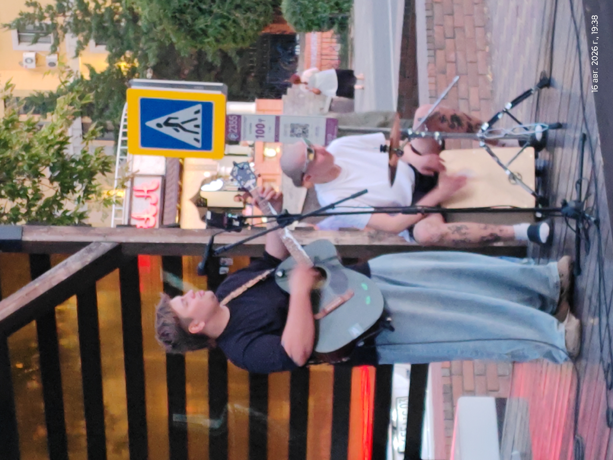 A man with short, messy brown hair is playing an acoustic guitar and singing into a microphone. He is wearing a black t-shirt and loose, light blue pants. He is standing on a wooden stage with a backdrop of horizontal black and yellow stripes. To his right, another man with a shaved head, wearing sunglasses and a white t-shirt, is playing a drum. He has tattoos on his legs. In the background, there is a pedestrian crossing sign, a building, and trees. The scene appears to be outdoors, possibly at an event or concert. The timestamp "16 авг. 2026 г., 19:38" suggests the date and time.